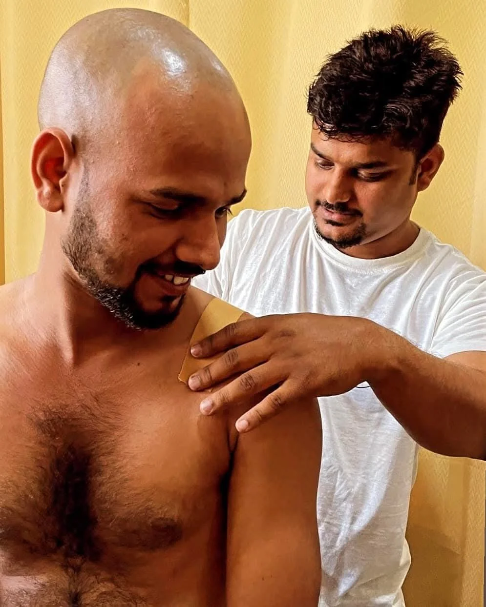 A medical professional is administering a vaccine injection to a shirtless man's upper arm, while the man smiles and looks down. The professional wears a white t-shirt and is focused on the injection. The background features a yellow wall.