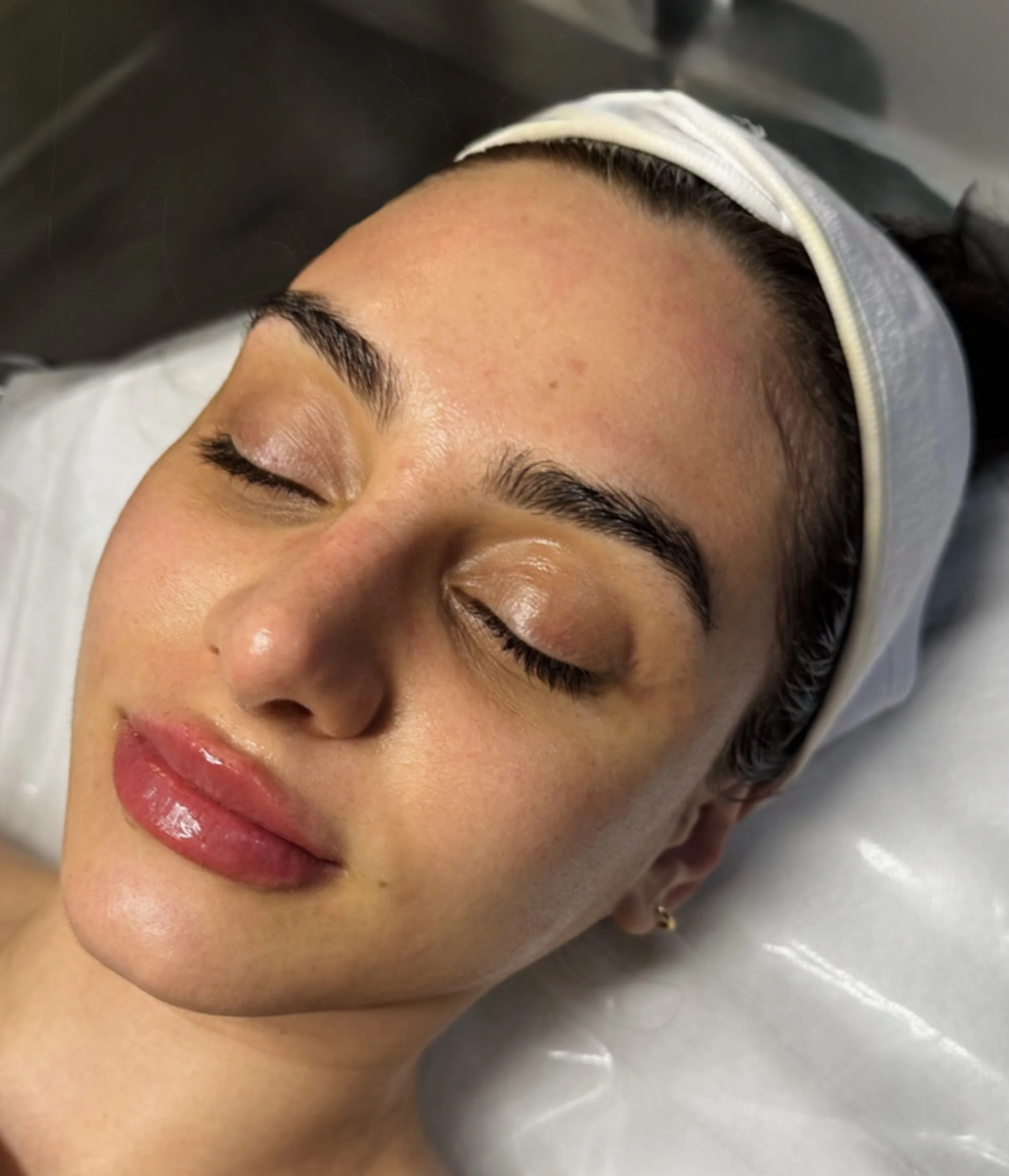 Close-up of a woman with clear skin, closed eyes, and makeup, lying down with a white headband.