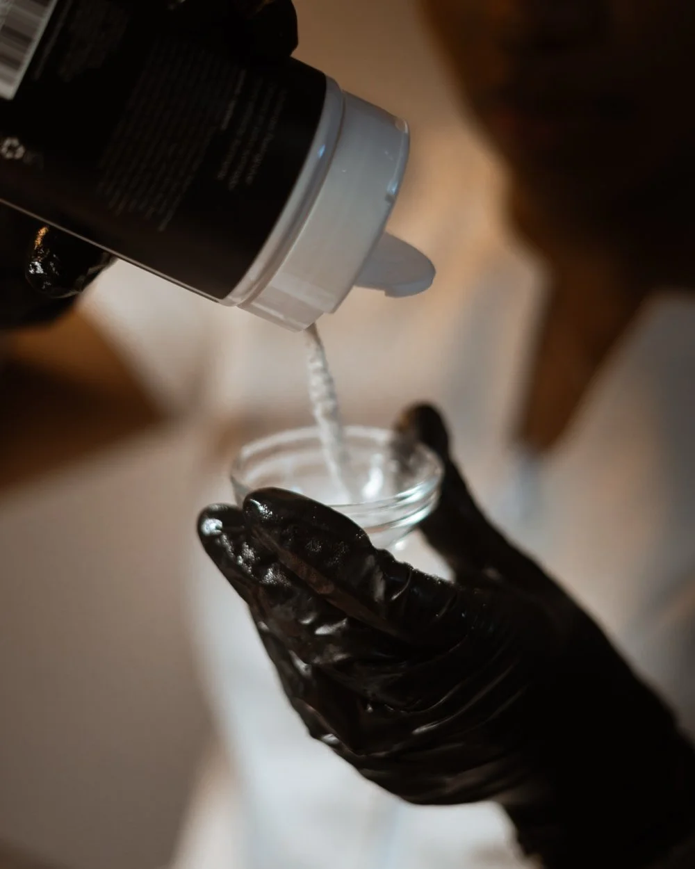 A hand wearing a black glove holding a small glass bowl as a liquid is poured into it from a bottle.