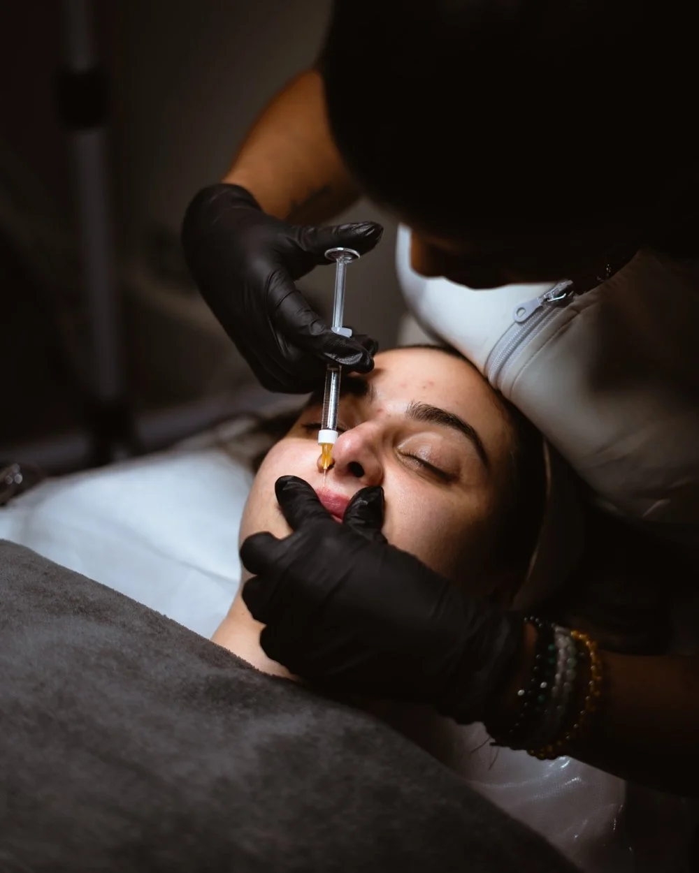 A person lying on a treatment bed receiving a nose filler injection from Camila Firetto