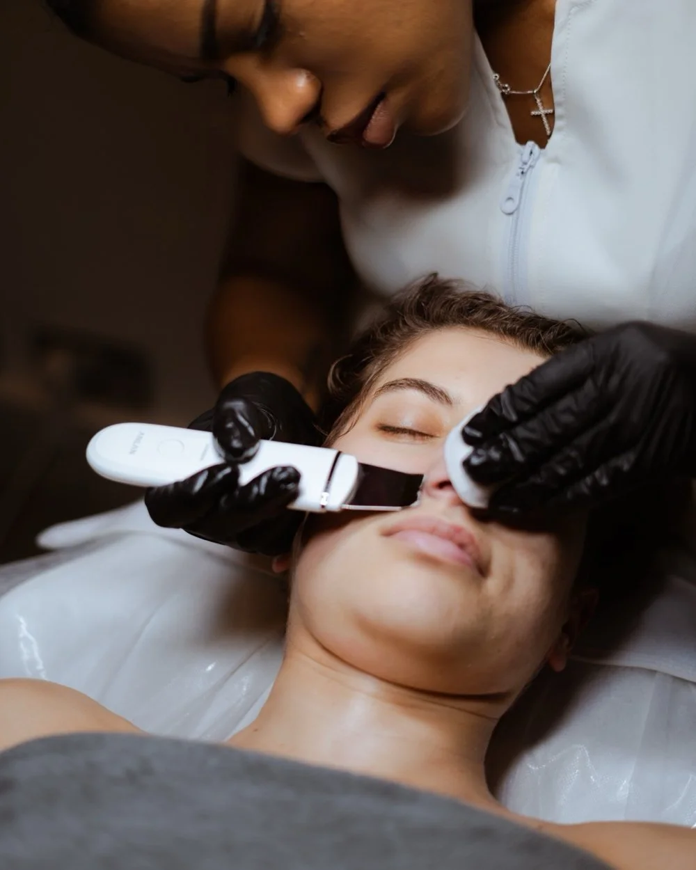 A woman receives a cosmetic facial treatment from a professional wearing black gloves, using a handheld device near her nose as she rests with eyes closed.