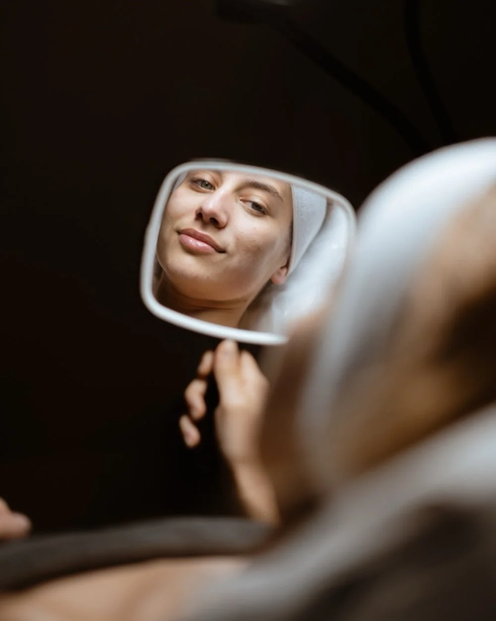 A woman with a towel on her head looking into a handheld mirror, looking at her lip filler