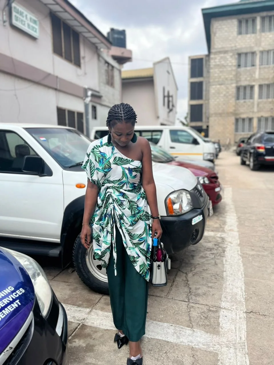 A woman in a tropical print wrap dress with one shoulder exposed, green pants, black and white shoes, carrying a black and white handbag, standing in a parking lot near several parked cars in an urban area with buildings and cloudy sky in the background.