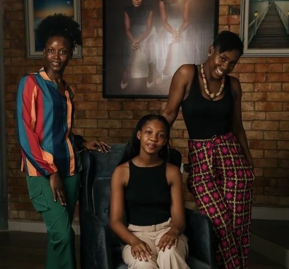 Three women in a cozy room with brick walls and framed artwork, posing for a photo. The woman on the left wears a multicolored striped shirt and green pants. The woman in the middle, seated, wears a black sleeveless top and beige pants. The woman on the right wears a black tank top, patterned pink and black pants, and a beaded necklace, smiling.