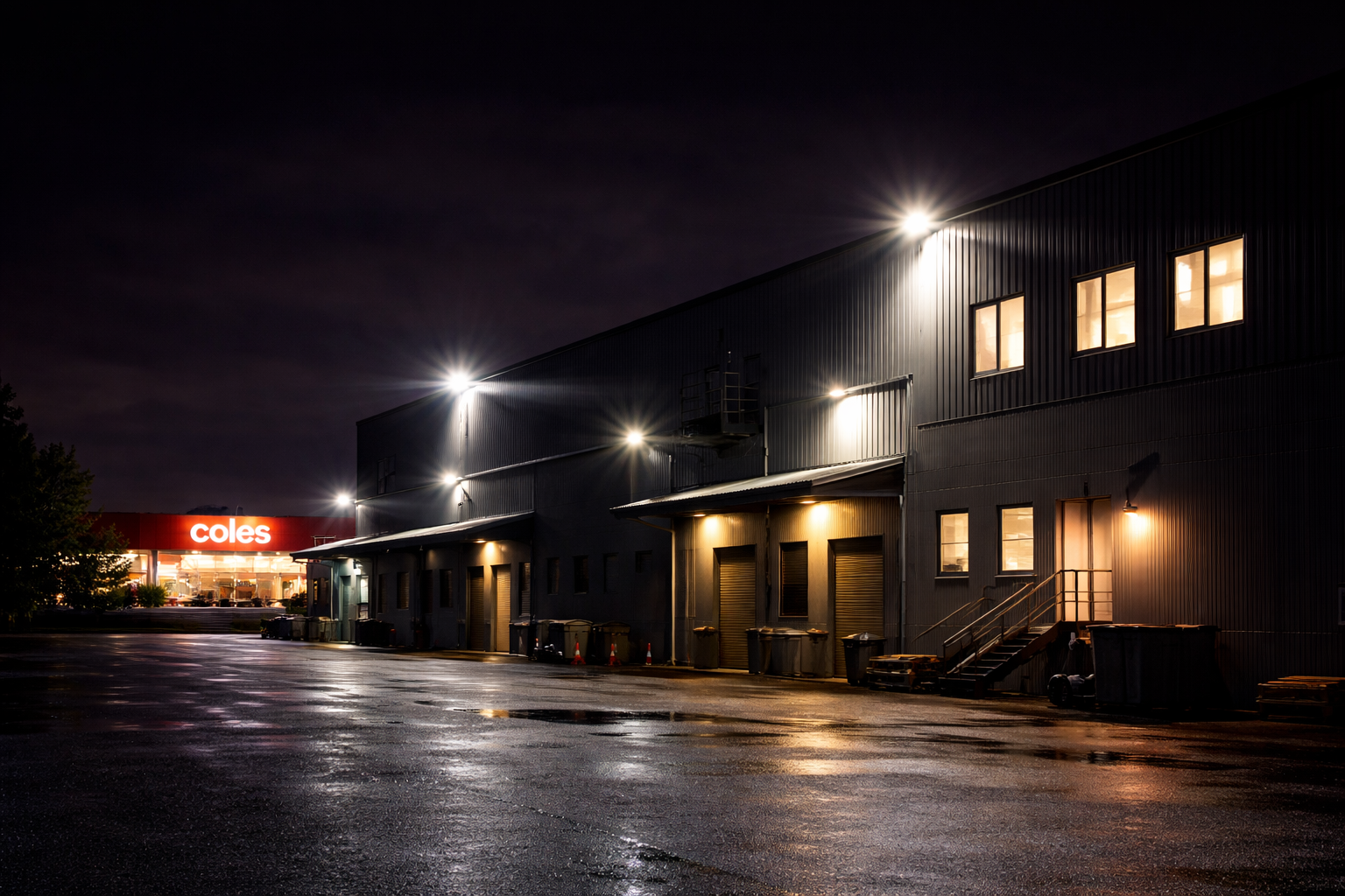 Nighttime view of a commercial building with illuminated windows and exterior lights, wet pavement reflecting light, and a distant store with a bright red sign that reads 'Coles'.
