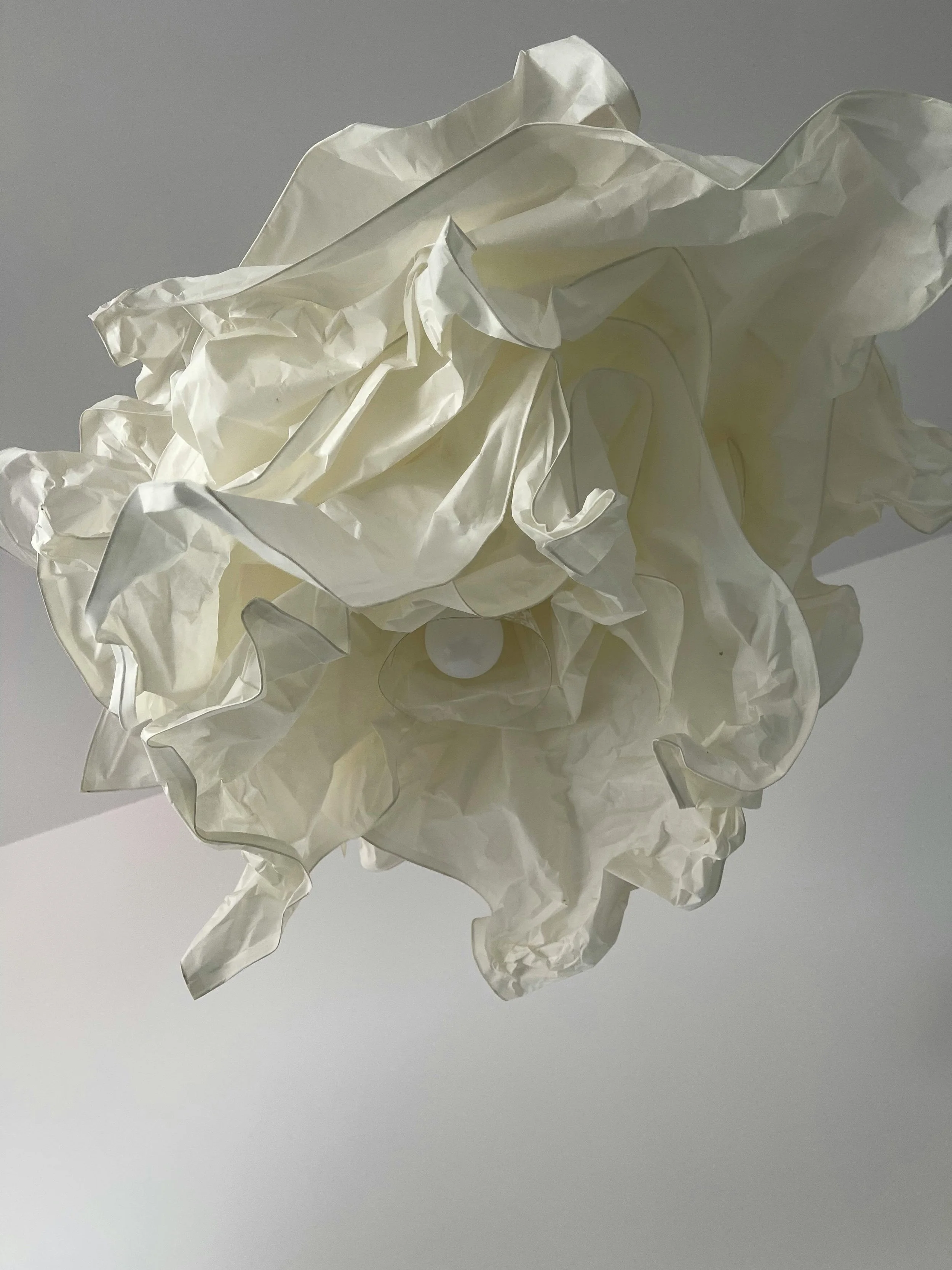 A crumpled white paper lamp shade with a light bulb inside, hanging from the ceiling against a plain background.