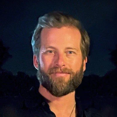 Man with a beard smiling, wearing a dark shirt, with a night background.
