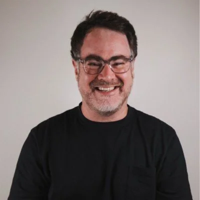 A man wearing glasses and a black shirt smiling against a plain background.