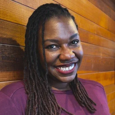 Smiling woman with long braided hair standing against a wooden wall.