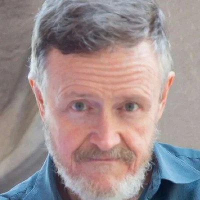 Portrait of an older man with short gray hair and a beard, wearing a blue shirt, with a neutral expression.