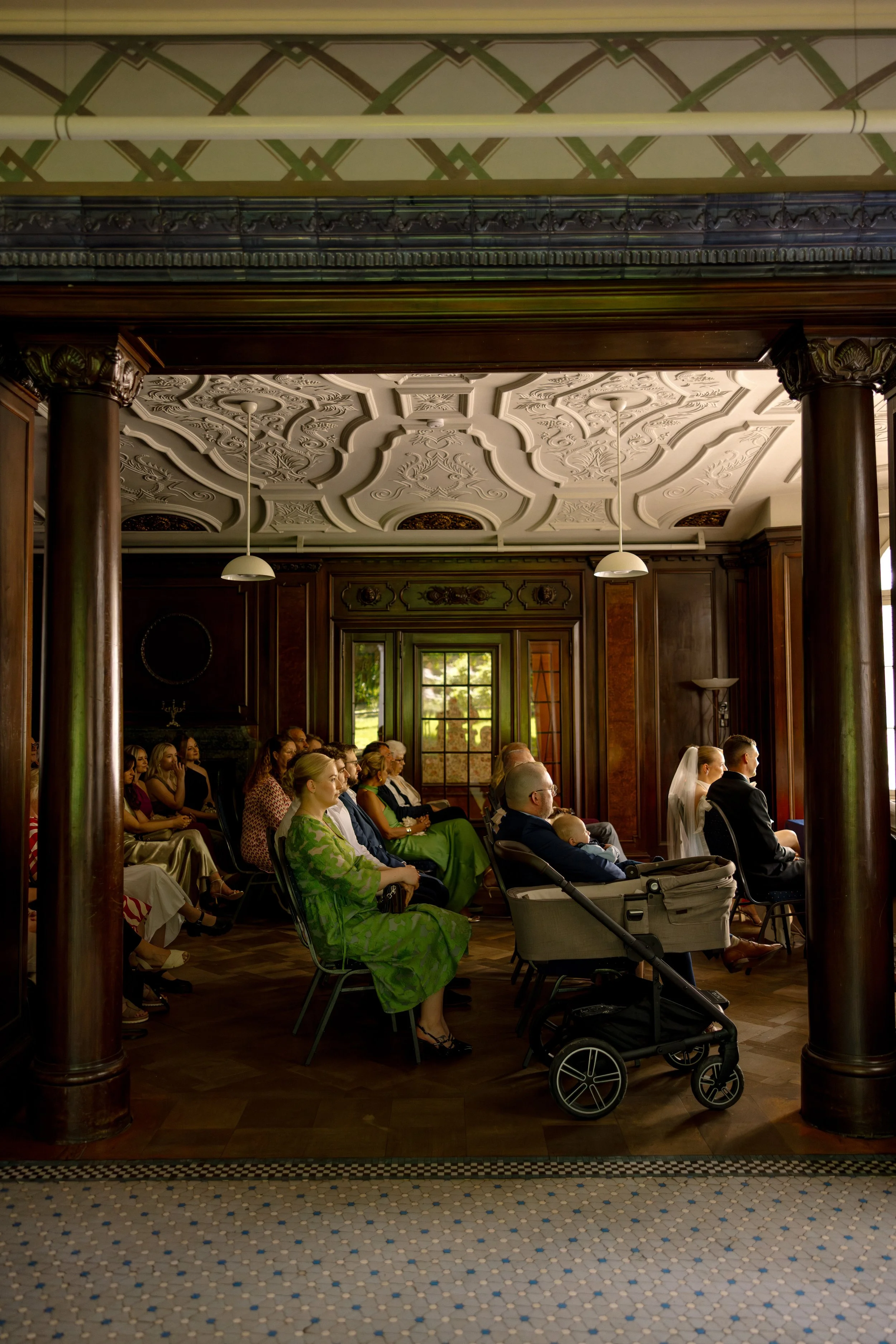 People seated indoors attending an event, with ornate ceiling and wood-paneled walls in the background.