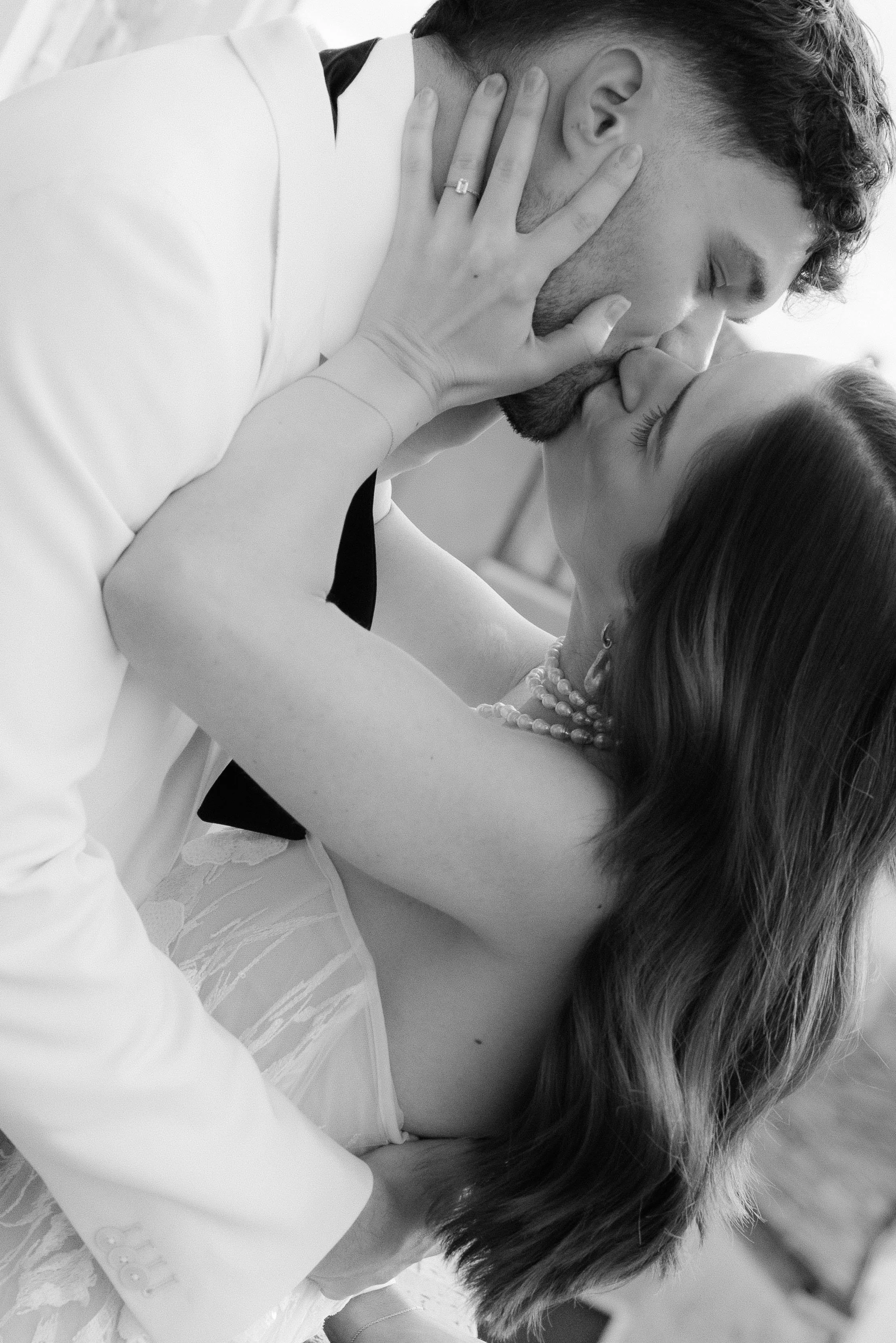A black and white photograph of a man and woman sharing a kiss, with the woman gently holding the man's face and wearing a pearl necklace.