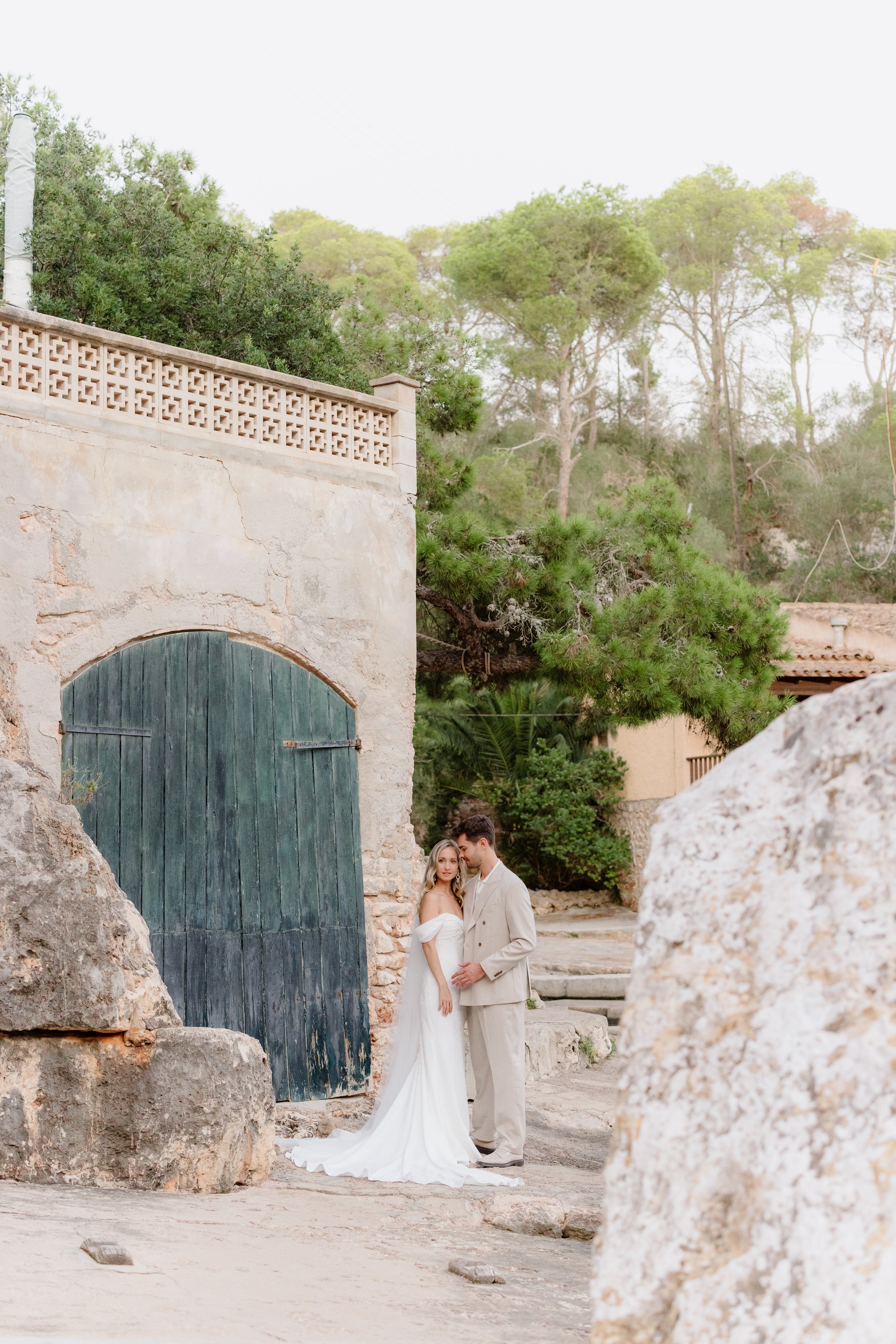 A bride and groom embracing outdoors near an old stone building with a large blue wooden door, surrounded by rocks, trees, and natural scenery.