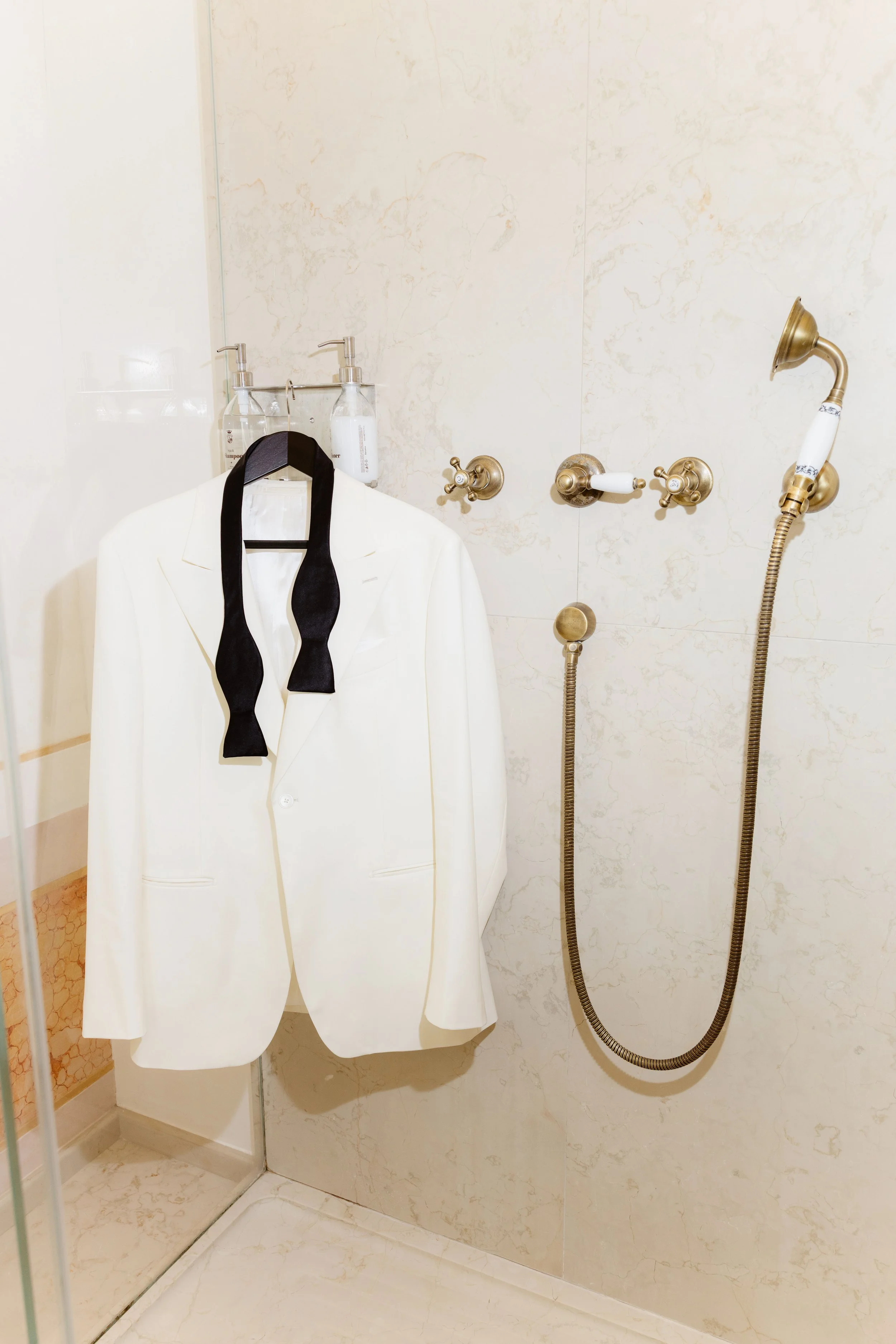 A bathroom shower area with a white blazer hanging on a black hanger, a black tie around its collar, and a marble-tiled wall with brass shower fixtures