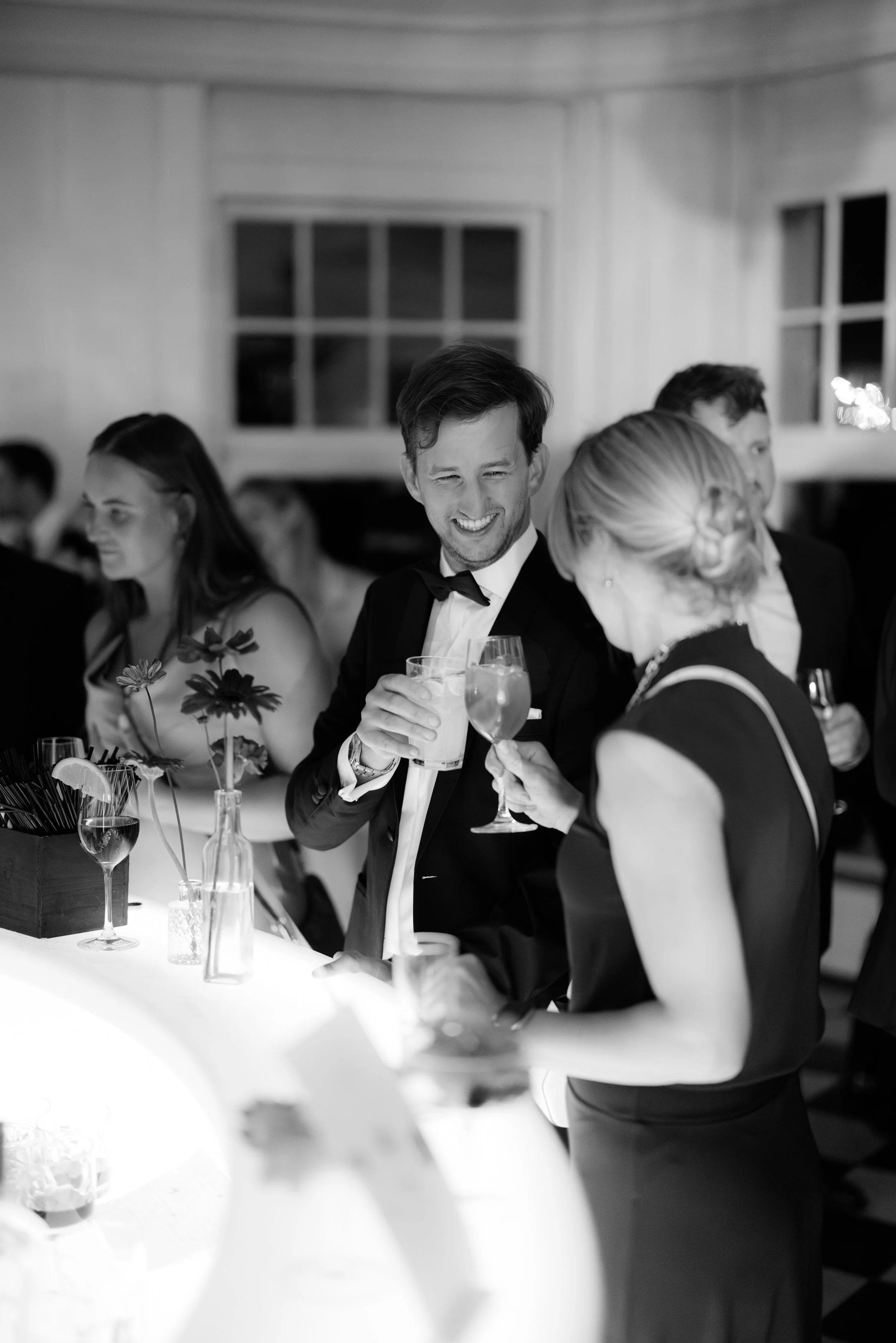 A group of people at a formal event, including a man in a tuxedo smiling and holding a drink, engaging in conversation with a woman who is also holding a drink, in a well-lit room with windows in the background.