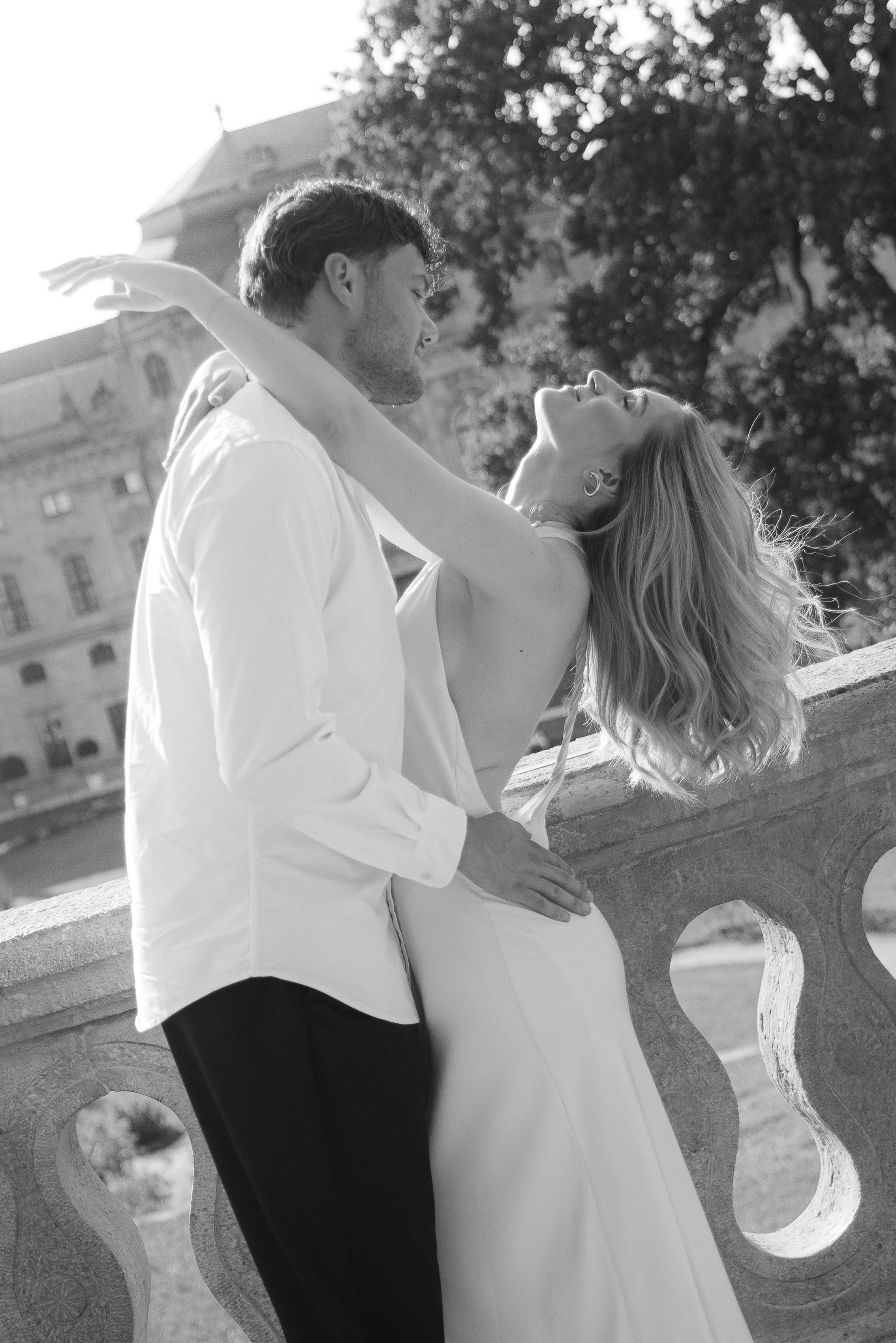 A black-and-white photo of a couple dancing outdoors on a bridge with a historic building and trees in the background. The woman has long, wavy hair and is leaning back with arms outstretched. The man is wearing a white shirt and dark pants, holding 