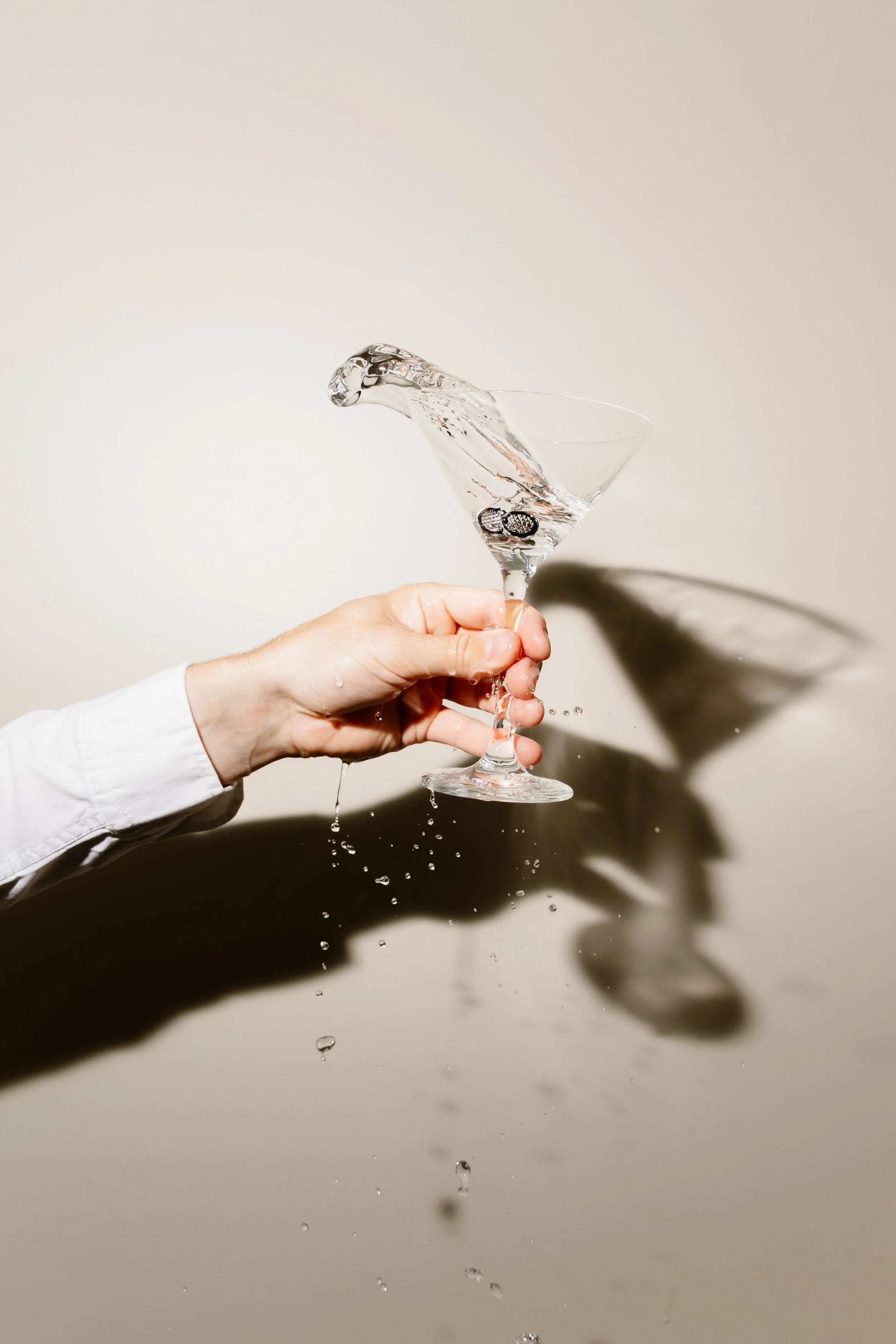 A hand holding a glass with water, causing water to splash out as it is poured, with a shadow cast on a plain background.