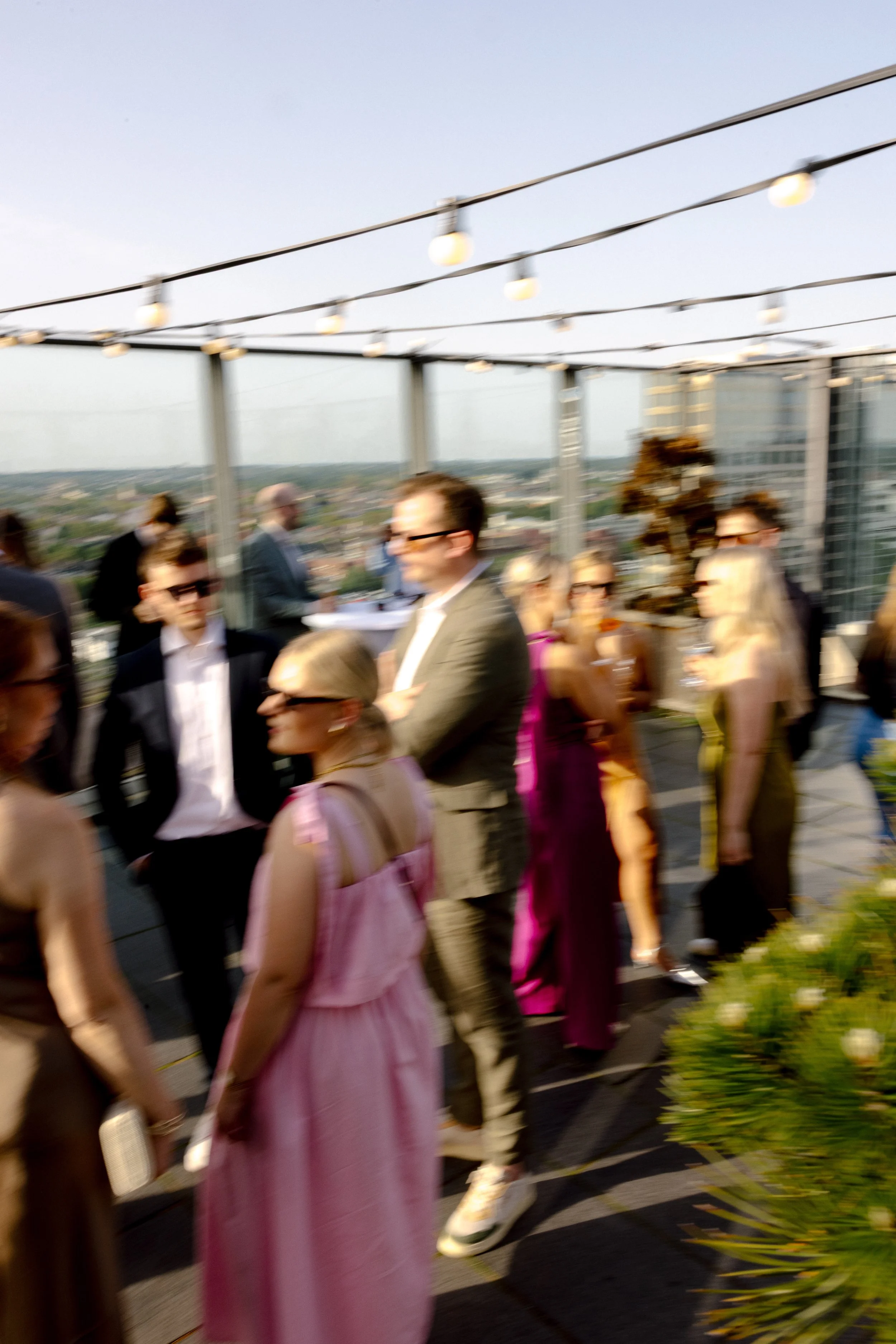 People dressed in formal and casual attire gathered on a rooftop terrace during sunset, with string lights hanging overhead and a cityscape in the background.