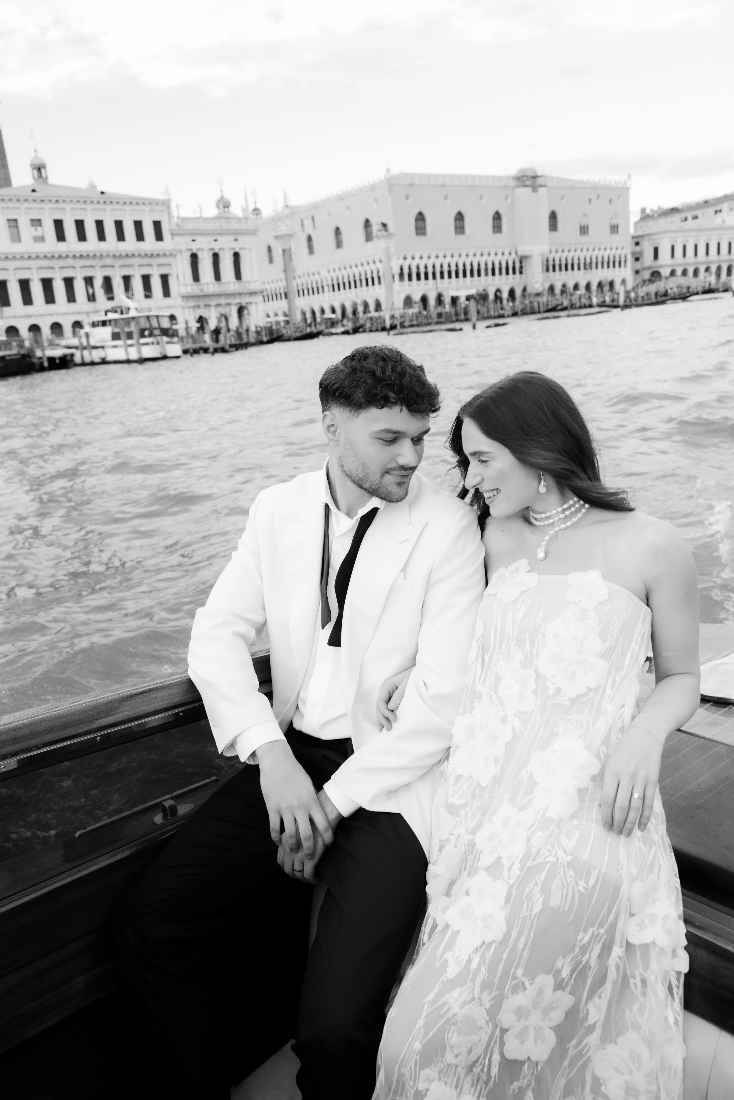 A couple dressed in wedding attire sitting on a boat, with a waterway and historical city buildings in the background.