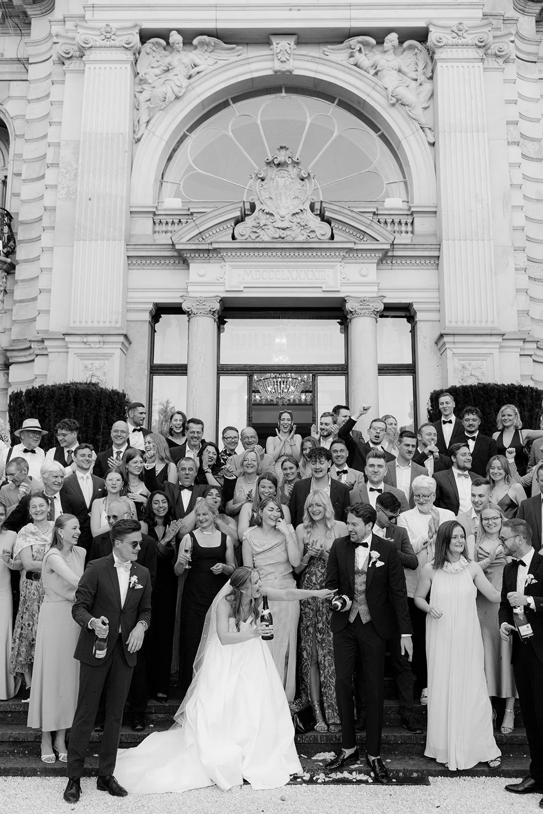 A large group of people dressed in formal clothes, gathered on the steps outside a grand building with ornate architecture, for a wedding celebration.