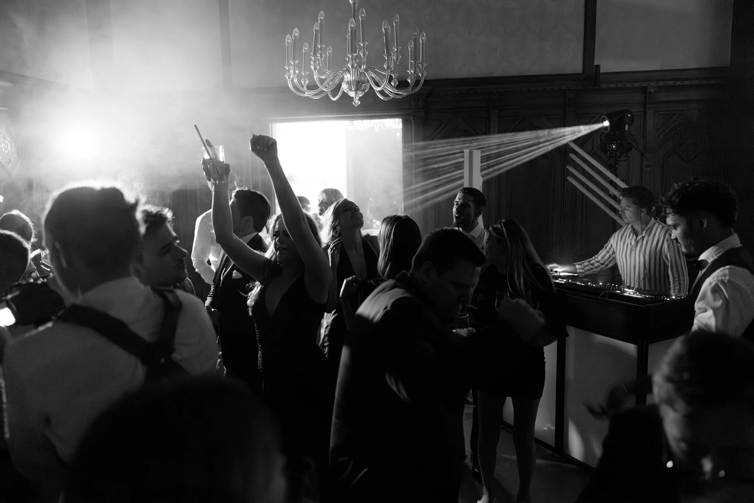 People dancing and enjoying music at a party or event with a DJ playing music, a chandelier, and beams of light.