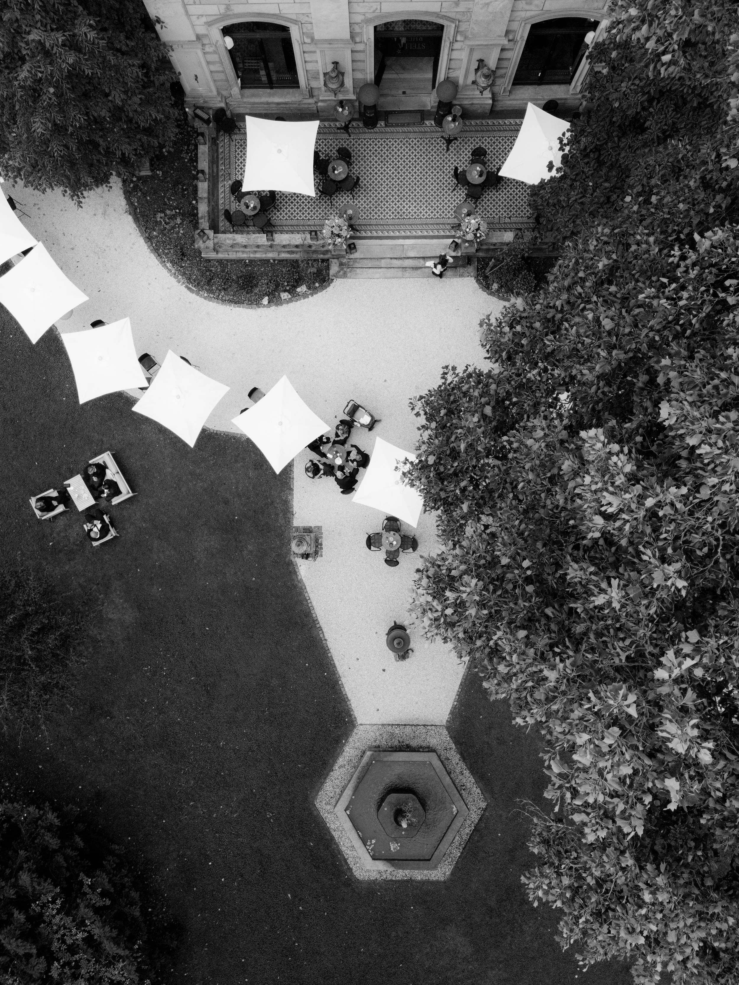 An aerial black-and-white photograph of an outdoor courtyard with a fountain, several tables with umbrellas, and trees surrounding the area.
