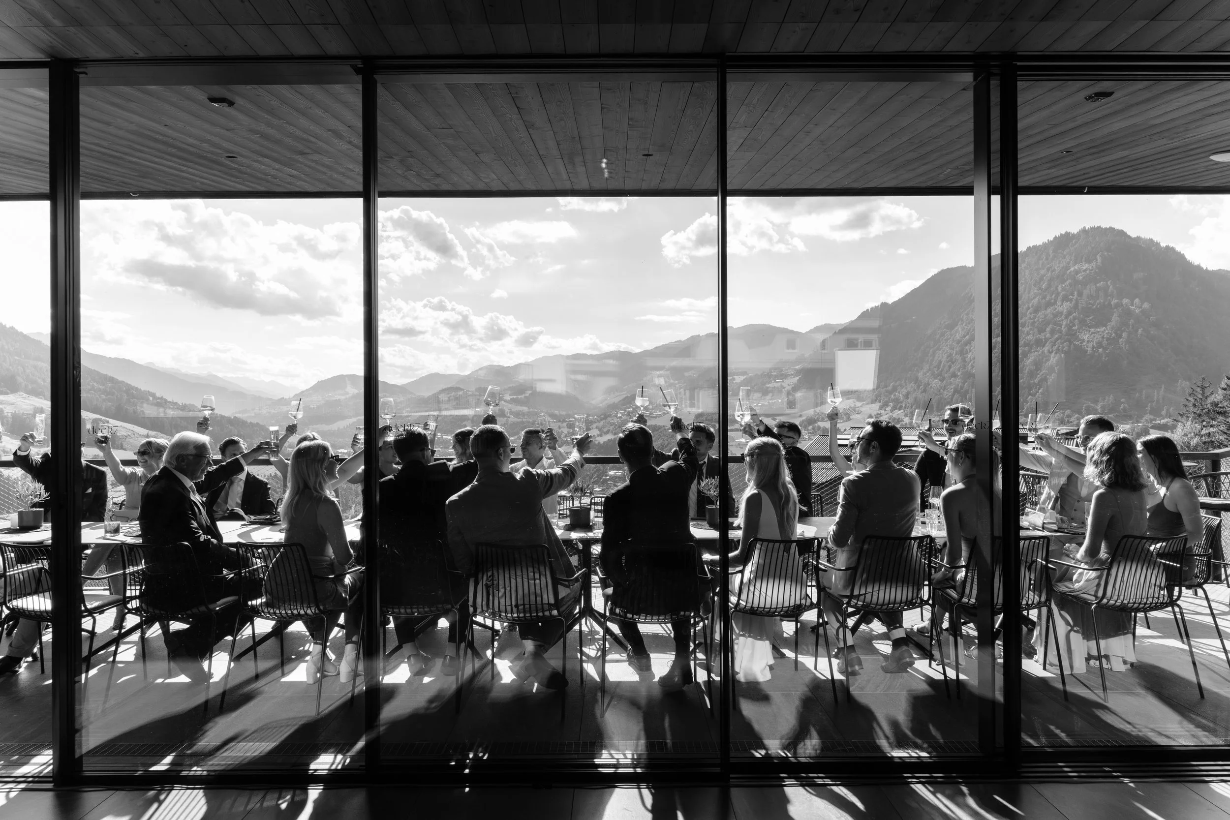 A group of people at a gathering or celebration, seated around a long table, raising glasses outdoors with mountain scenery in the background, seen through large glass windows.