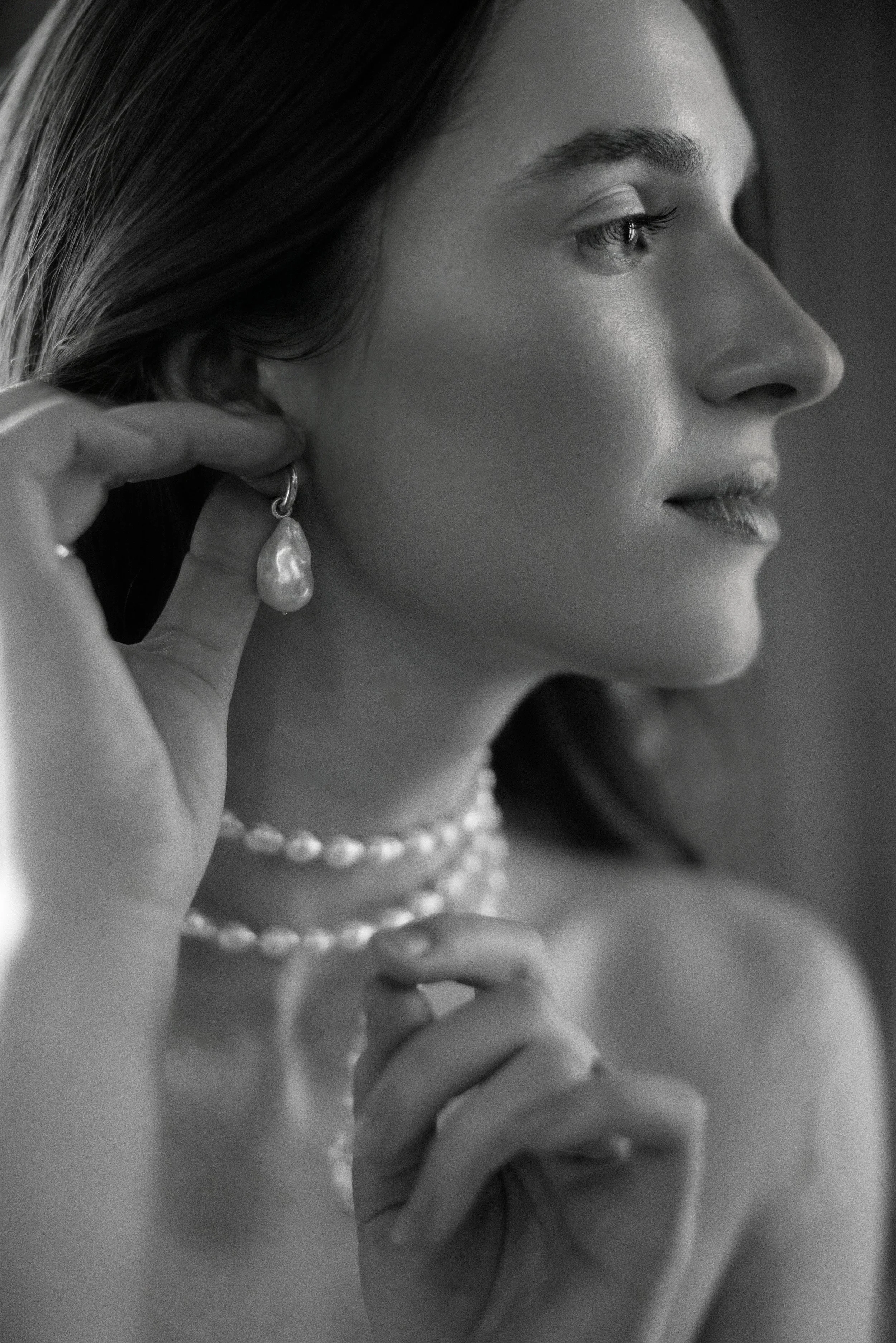 Black and white photograph of a woman wearing pearl jewelry, including earrings, necklace, and a ring, with her hand near her neck and her head slightly turned to the side.