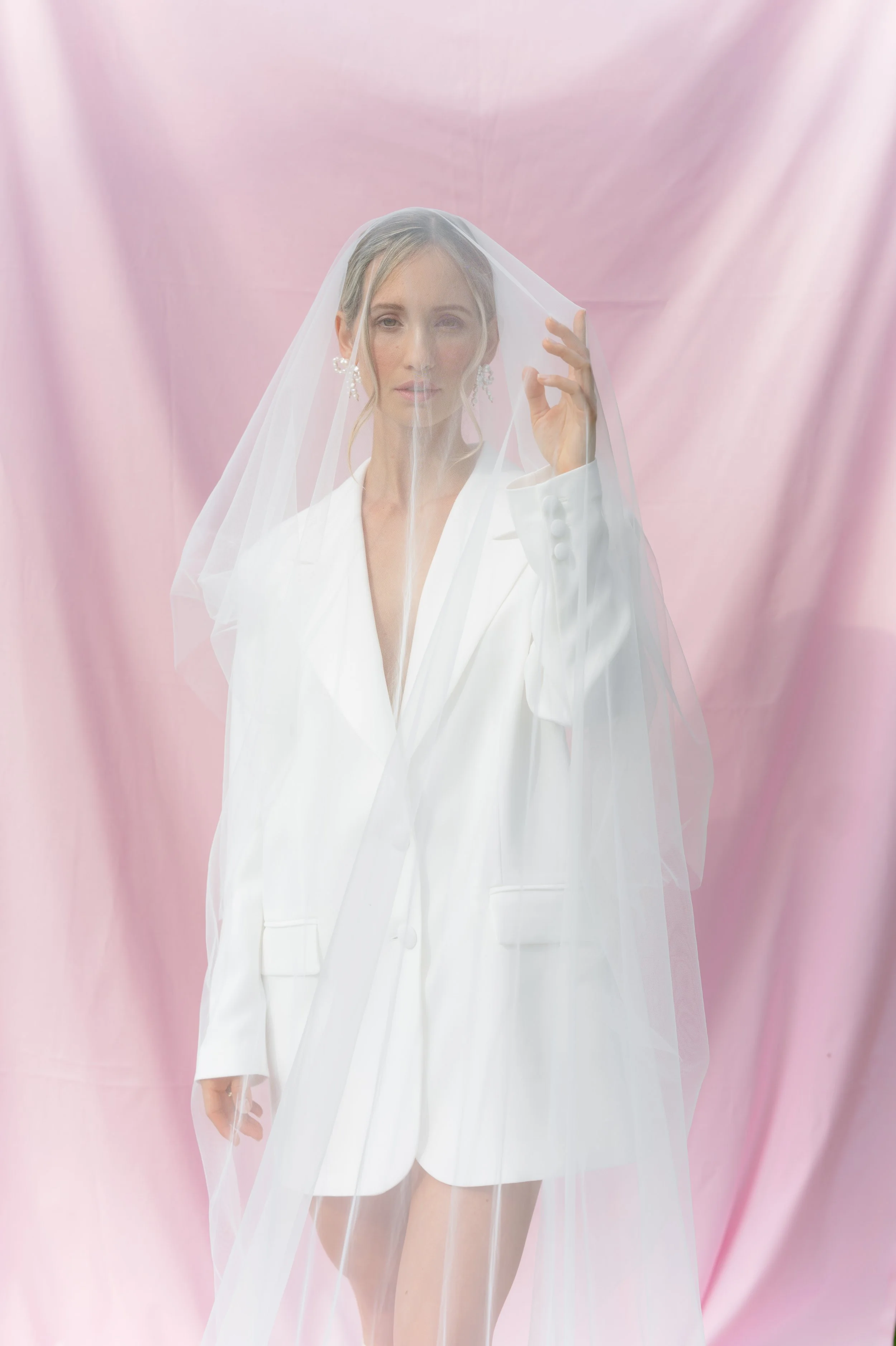 A woman dressed in a white blazer and matching shorts, wearing a veil, standing in front of a pink background.