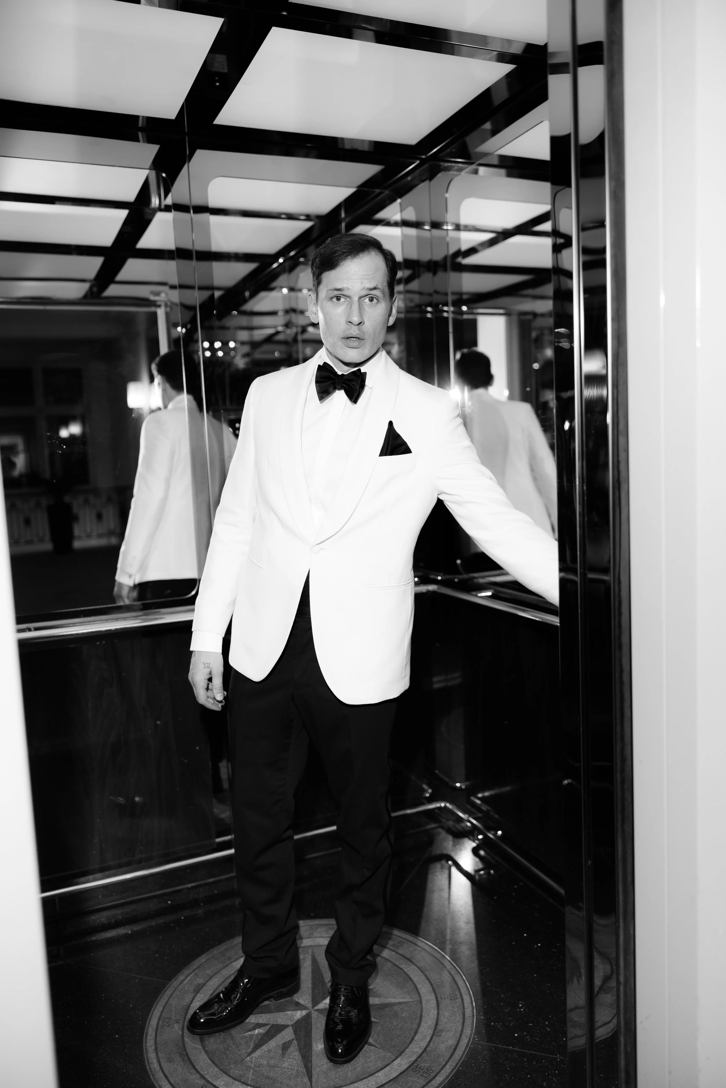 Man wearing a white tuxedo jacket, black bow tie, and black pants inside an elevator with mirrored walls.
