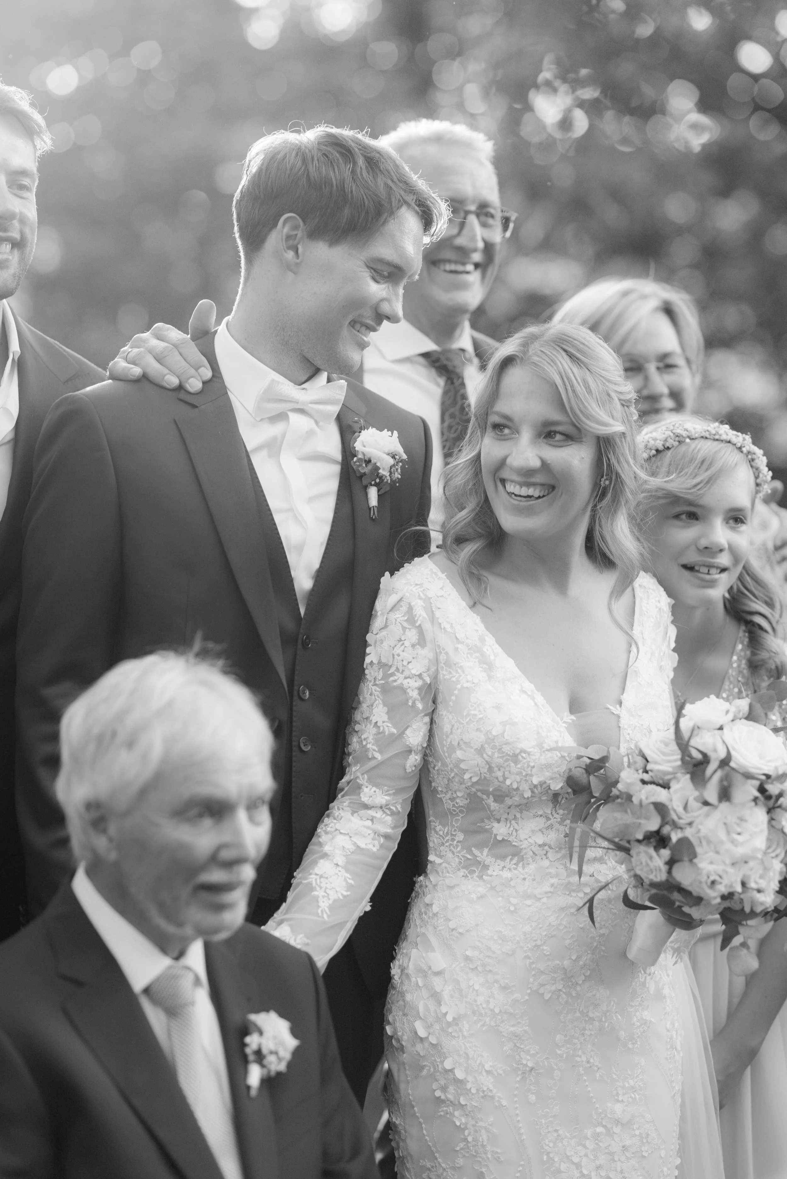 Black and white photo of a wedding celebration with a bride in a lace gown holding a bouquet, and groom in a tuxedo, smiling at each other, surrounded by family and friends.