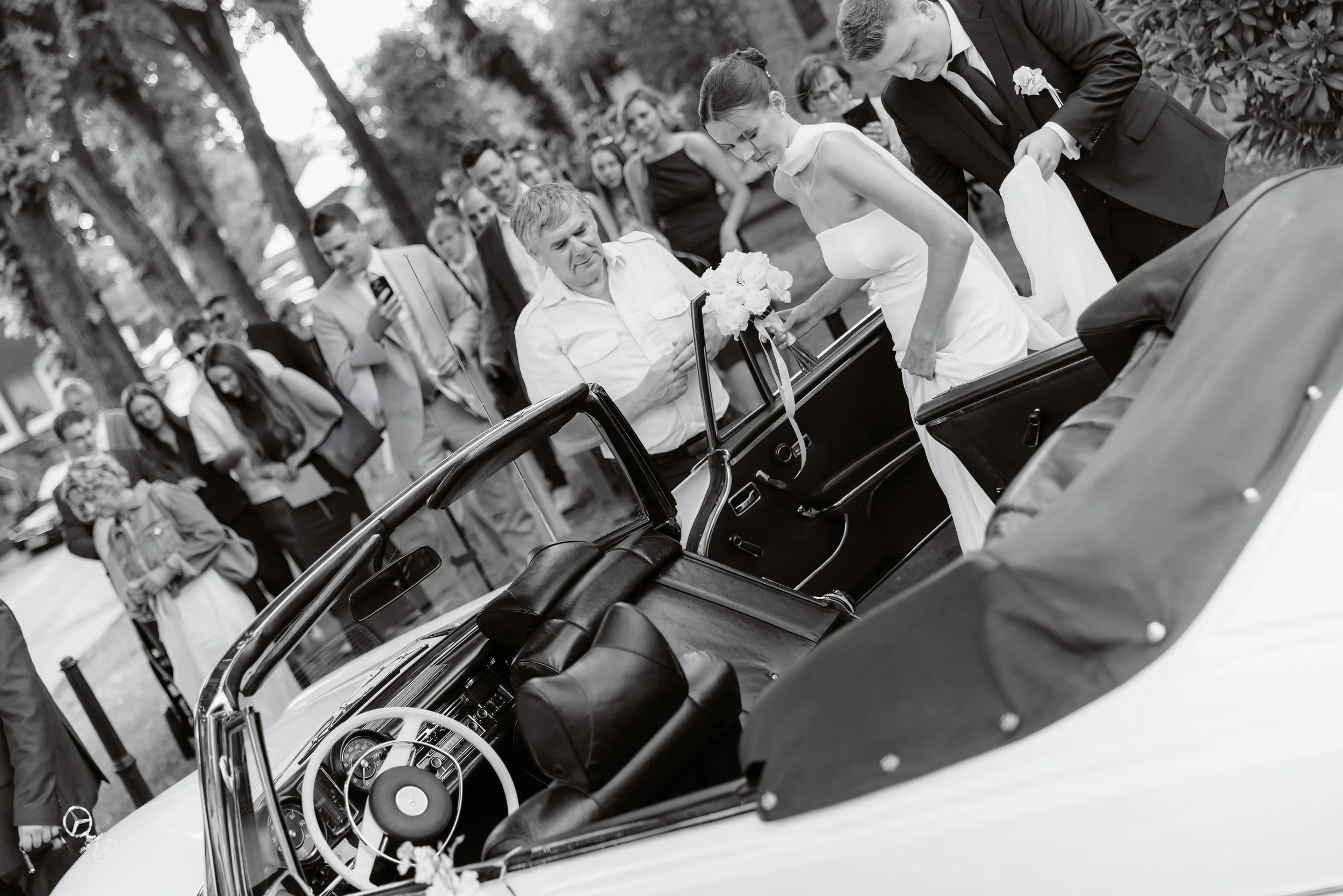 A bride in a white wedding dress gets out of a vintage car on a street lined with trees during a wedding celebration, with guests and family in the background.