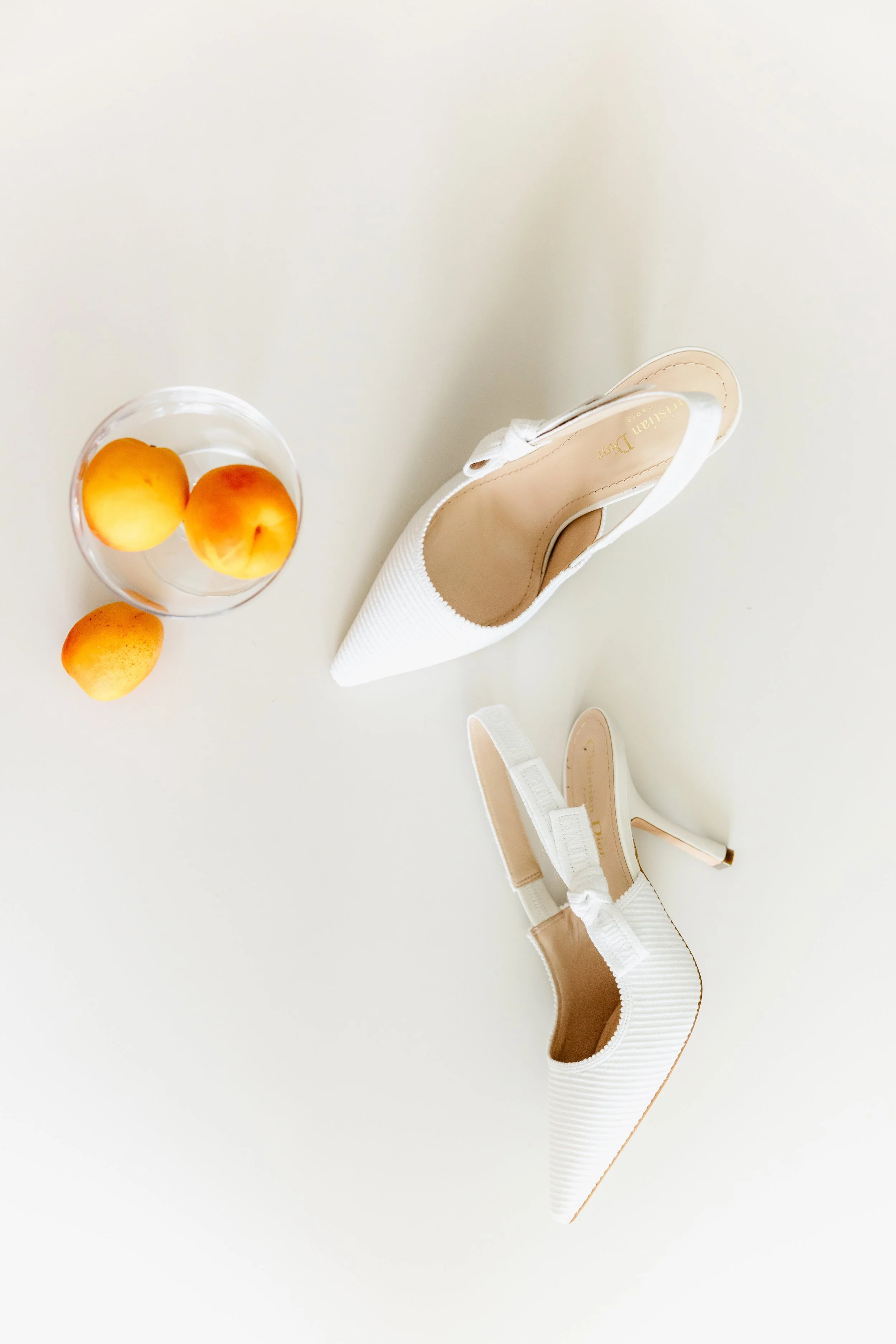 White high-heeled shoes and a glass bowl with three peaches on a white surface.