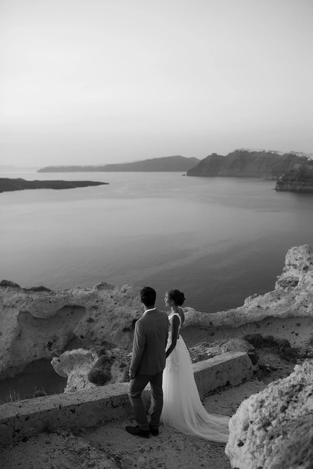 A couple dressed in wedding attire, standing on rocky terrain overlooking a calm body of water with cliffs in the distance, black and white photo.