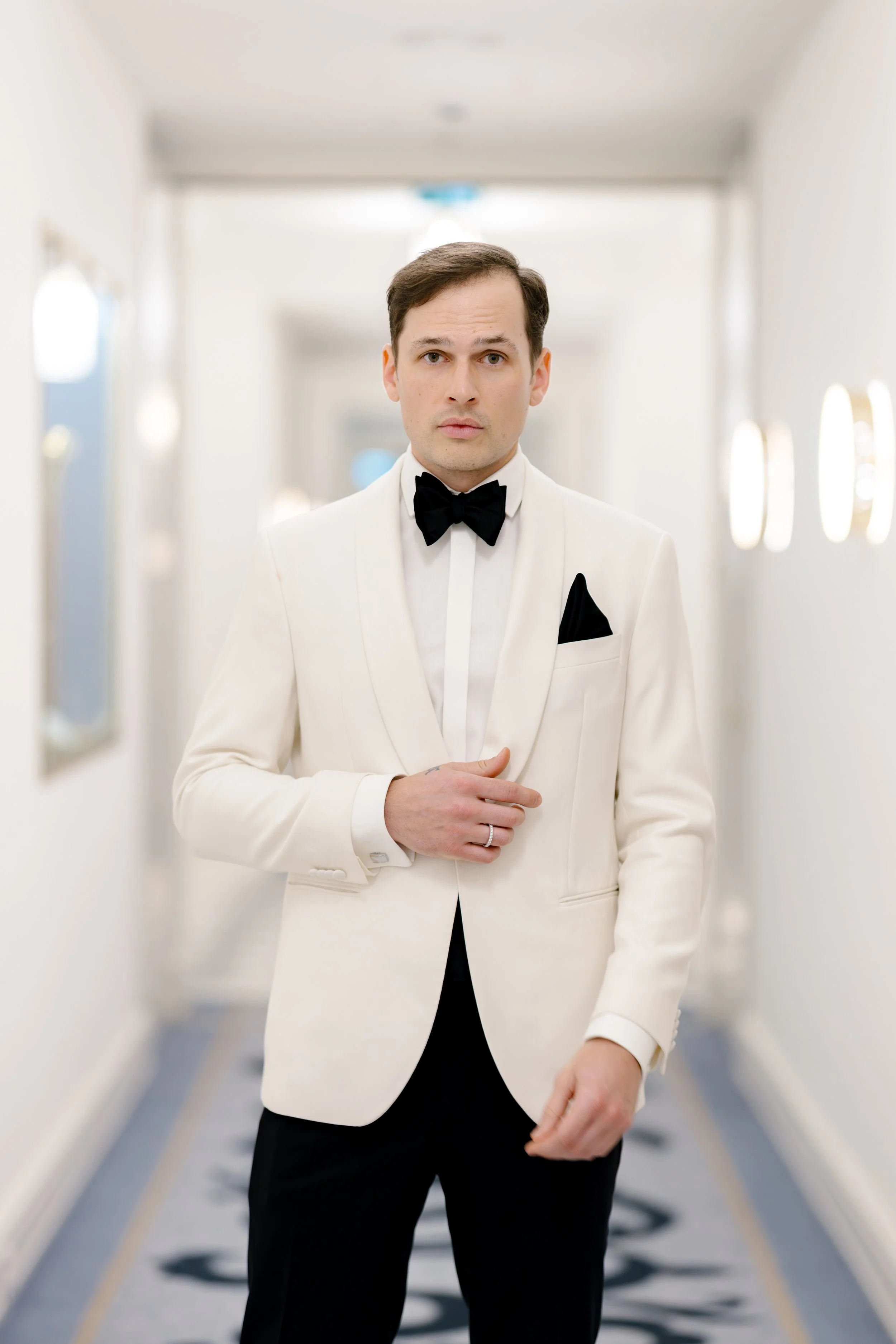 A man in a white tuxedo with a black bow tie and pocket square standing in a hotel corridor.