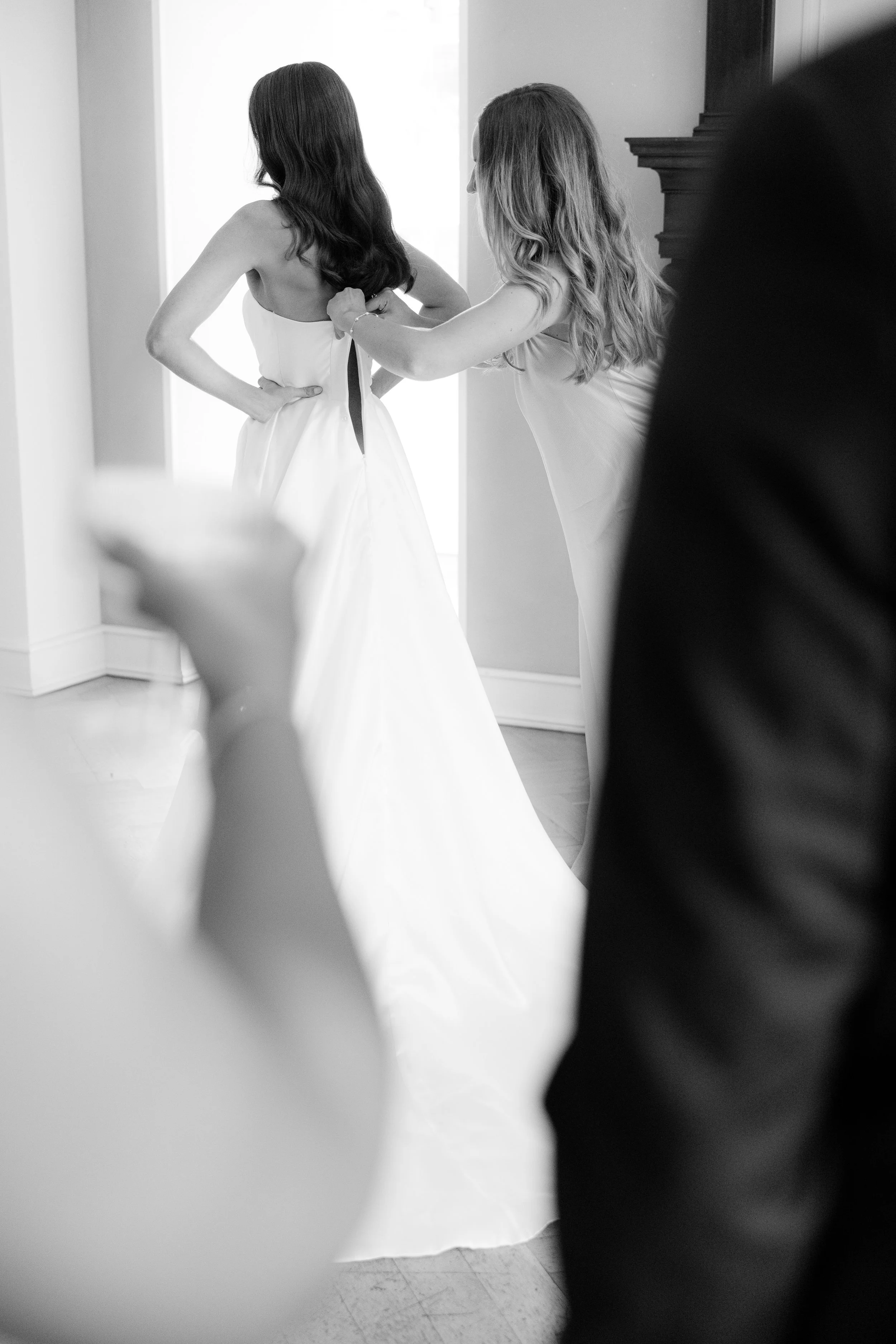 A bride is getting ready for her wedding, with a woman helping her fasten the back of her wedding dress in a room with natural light.