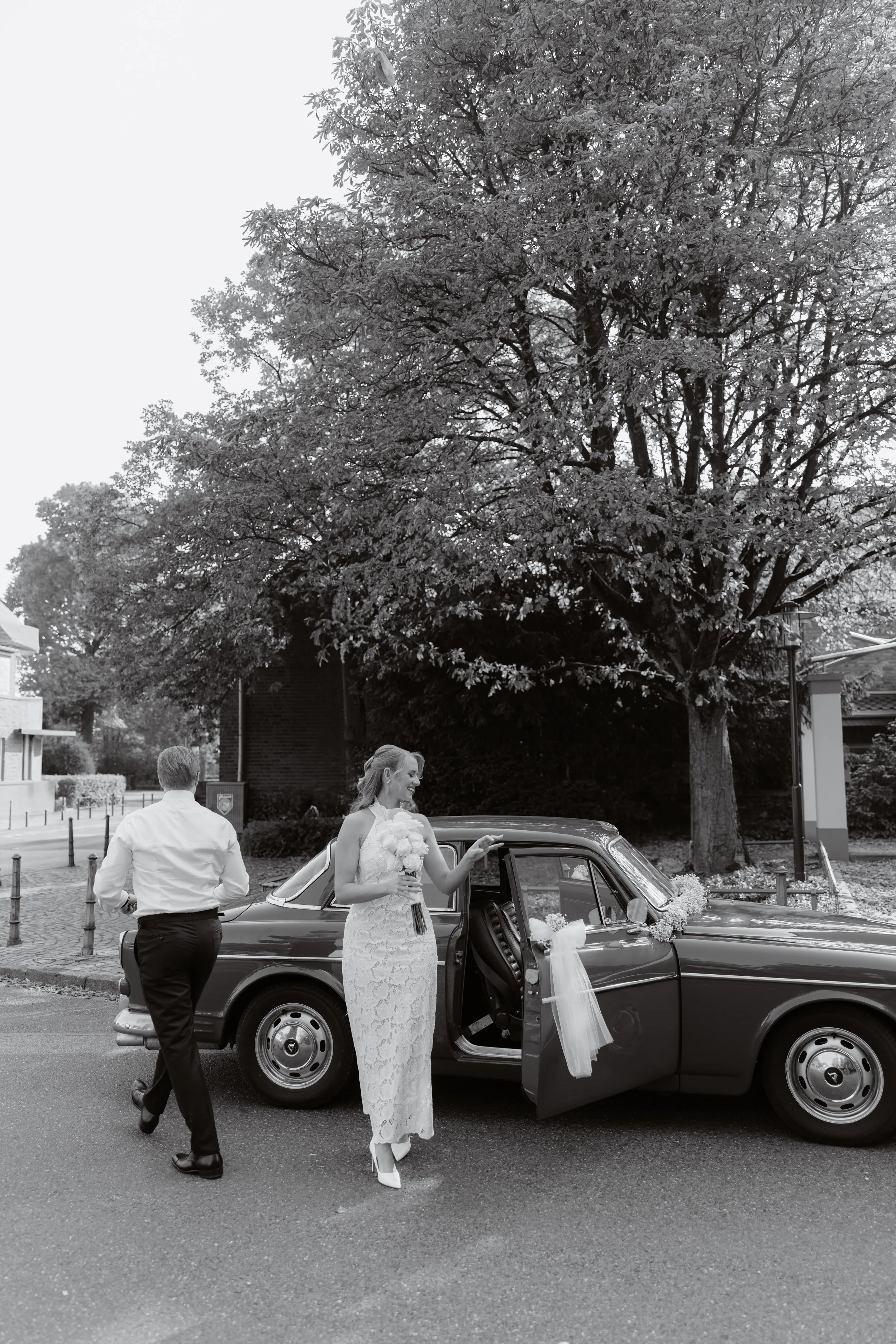 A bride and groom preparing to leave in a vintage car decorated with flowers. The bride is holding a bouquet and smiling, while the groom is walking towards the car on a street with large trees in the background.