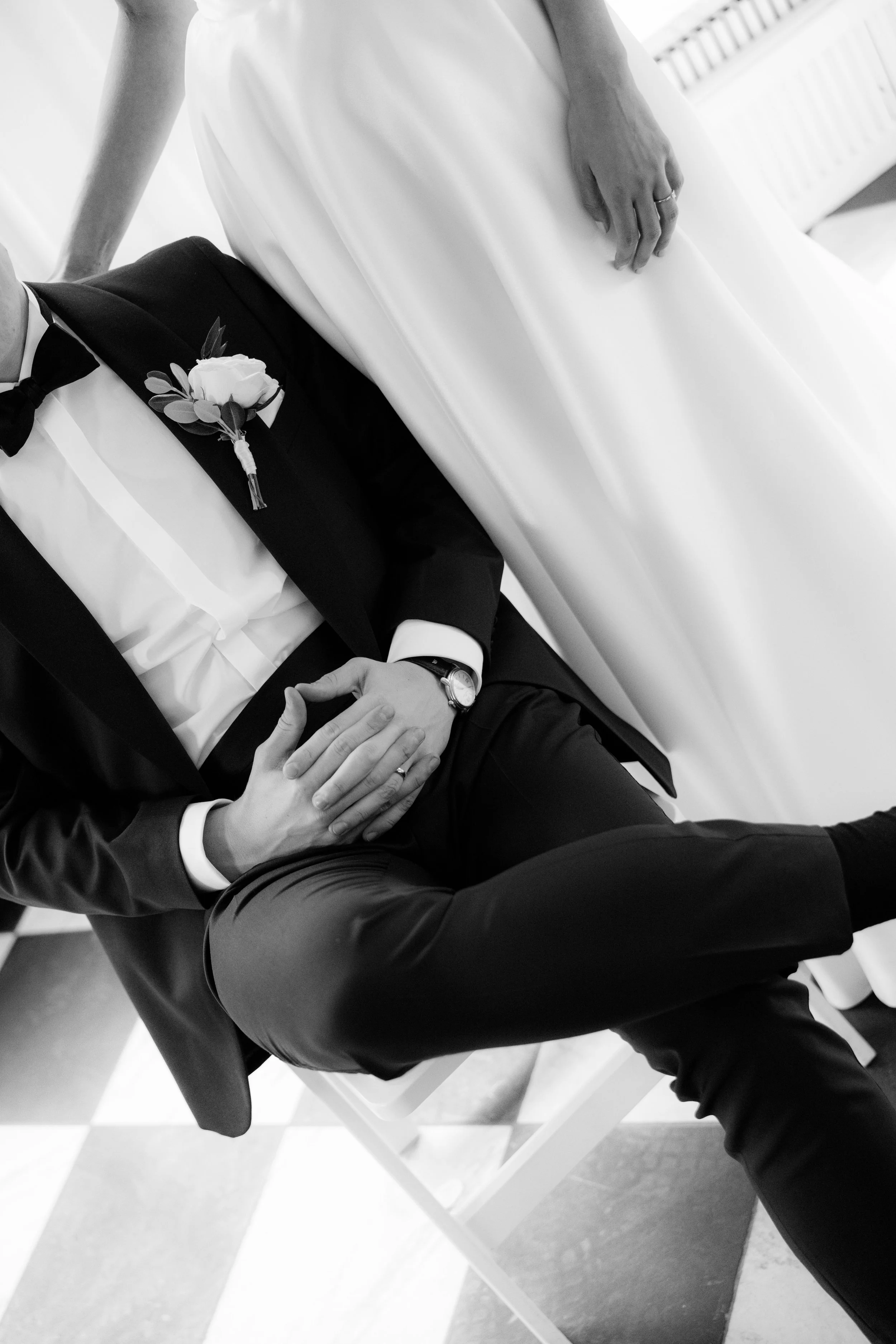 Black and white photo of a man in a tuxedo sitting on a chair, holding a woman's hand, with a visible wedding band. The woman, wearing a white dress, is standing next to him with her hand resting on his arm.