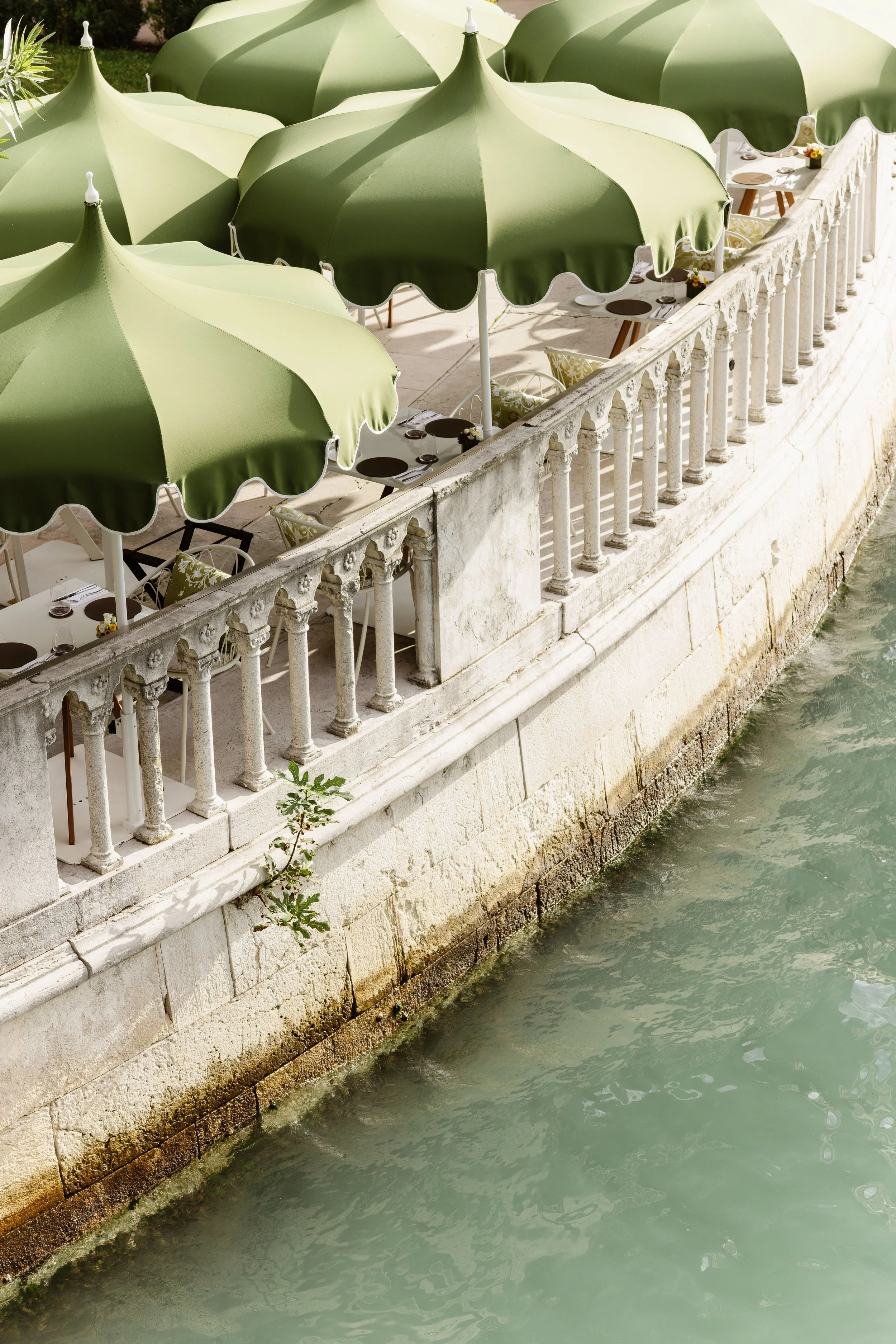 Outdoor seating area with green umbrellas along a stone waterfront railing.