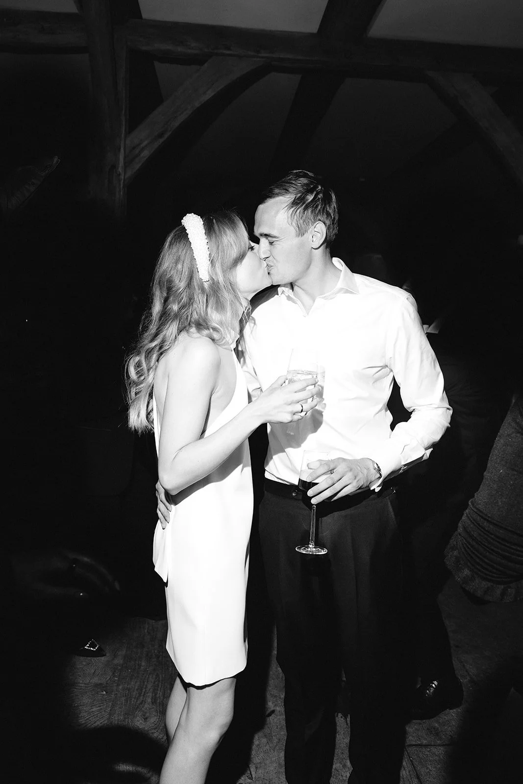 A black-and-white photo of a man and woman kissing, holding drinks, in a dimly lit indoor setting.