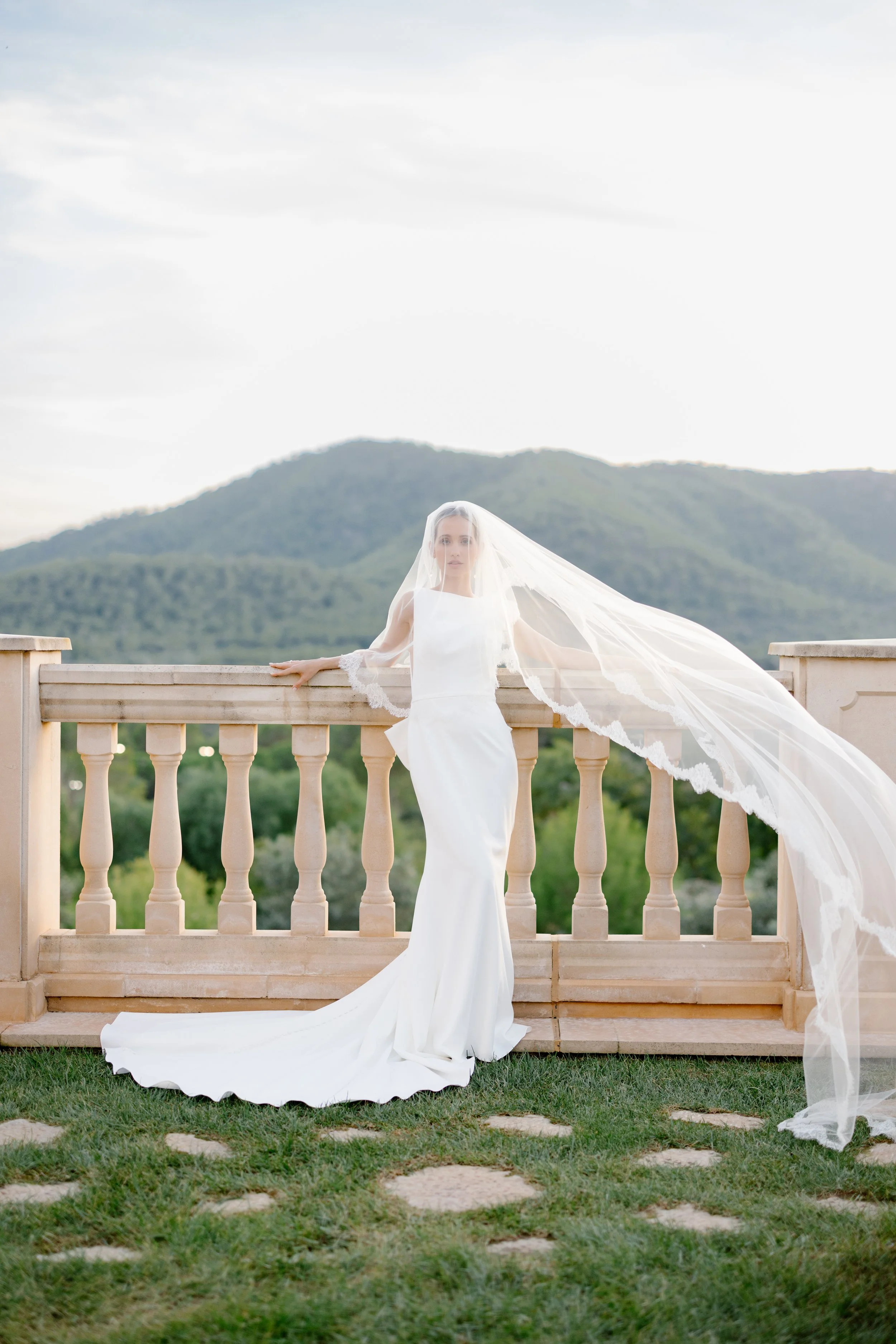 A bride in a white wedding dress with a long train and veil, standing on a stone-paved grass area in front of a stone railing with mountain scenery in the background.