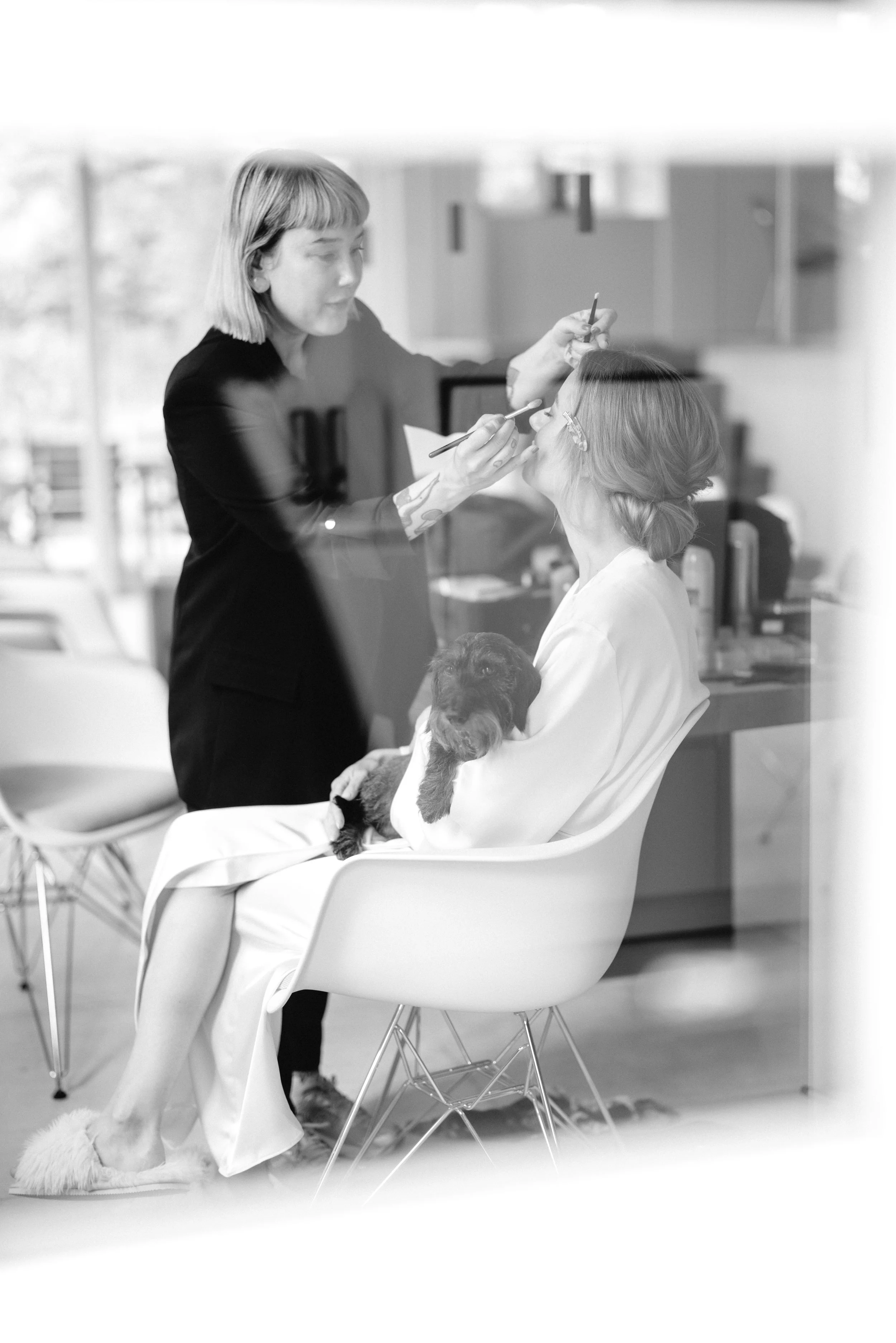 A woman sitting in an armchair getting her makeup done at a beauty salon, holding a small dog on her lap, with a stylist applying makeup and a window in the background.