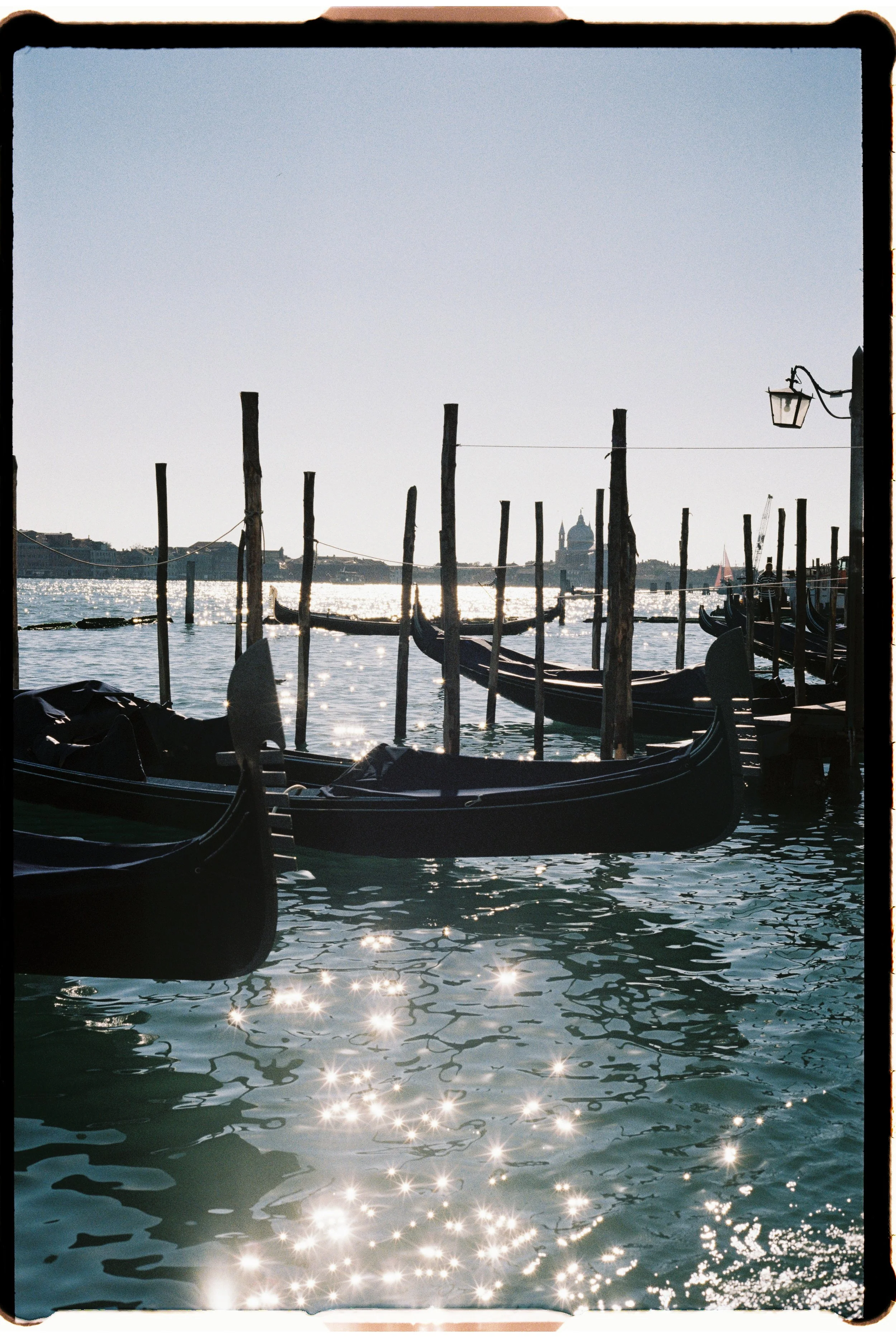 View of gondolas docked along a canal with sunlight reflecting off the water and historic buildings in the background, including a domed church or basilica.