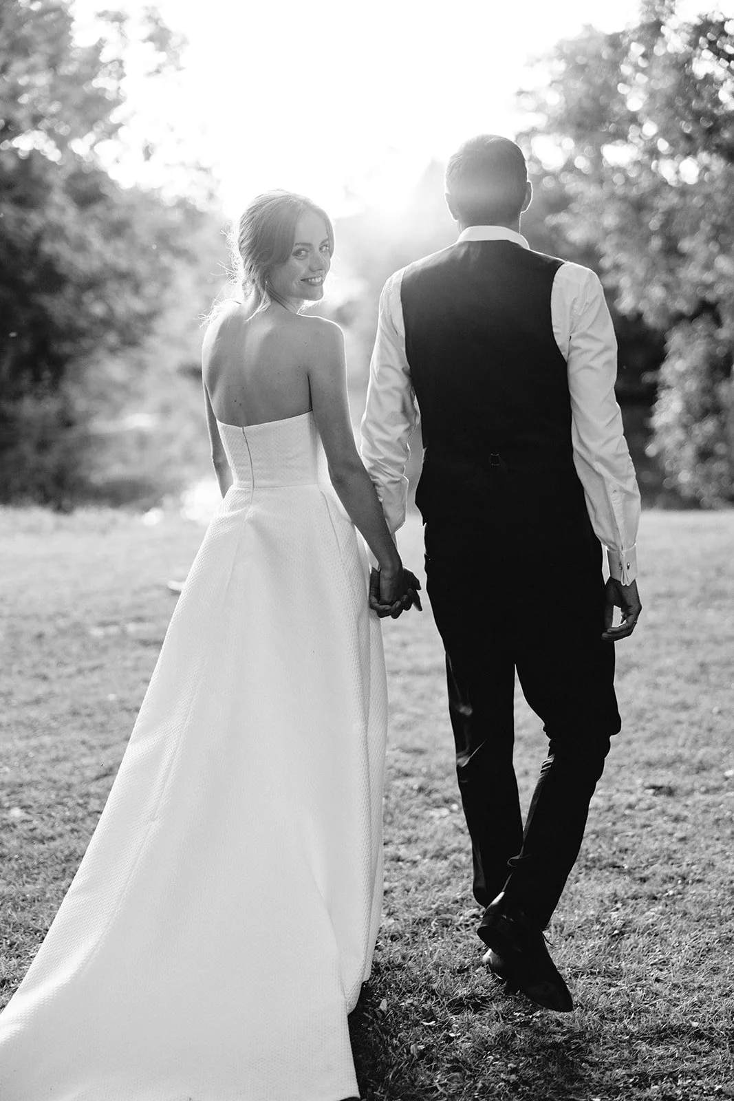 A bride and groom walking hand in hand outdoors during sunset, smiling back at the camera, black and white photograph.