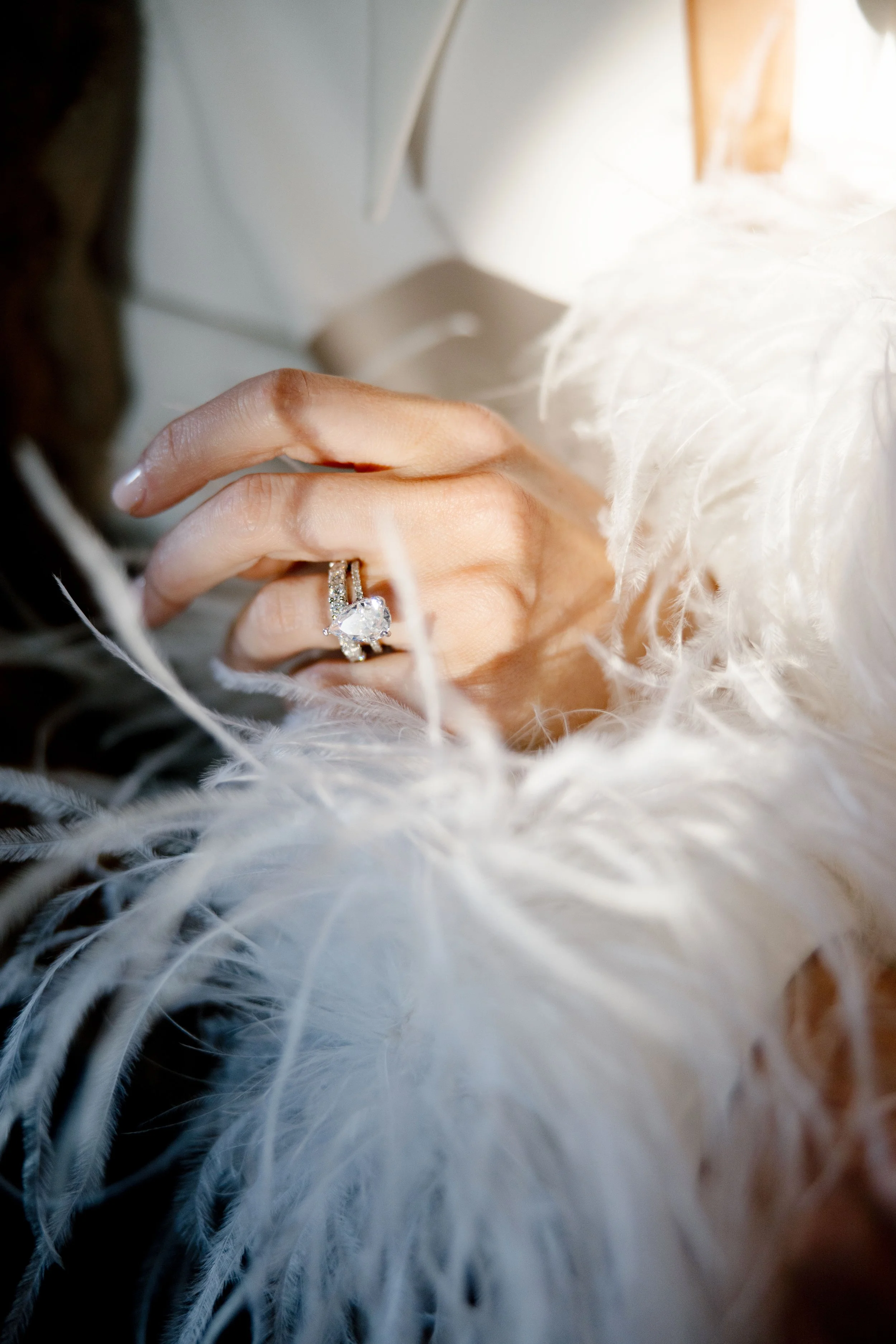 Close-up of a woman’s hand showing a large, sparkling diamond engagement ring, surrounded by white feathers.