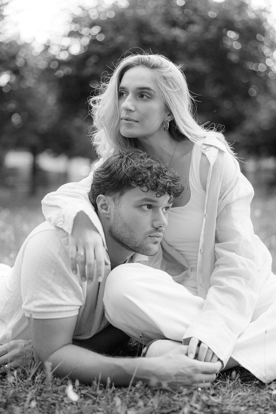 A black-and-white photo of a young woman with blonde hair and a young man with curly hair outdoors, with trees in the background. The woman is sitting behind the man, resting her arm over his shoulder, while the man is lying on the grass, supporting 