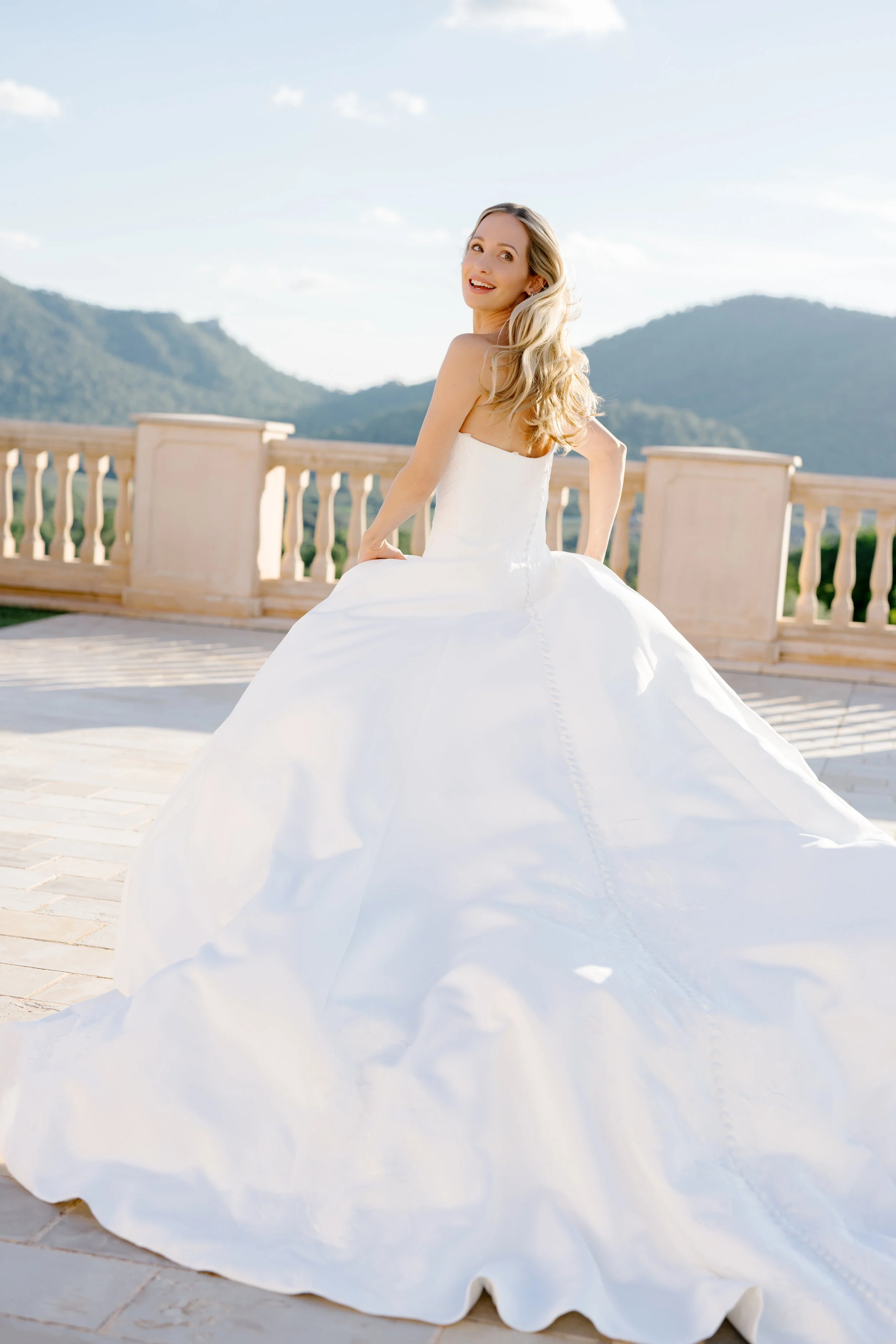 A woman in a white wedding dress smiling outdoors on a sunny day, with mountains in the background.