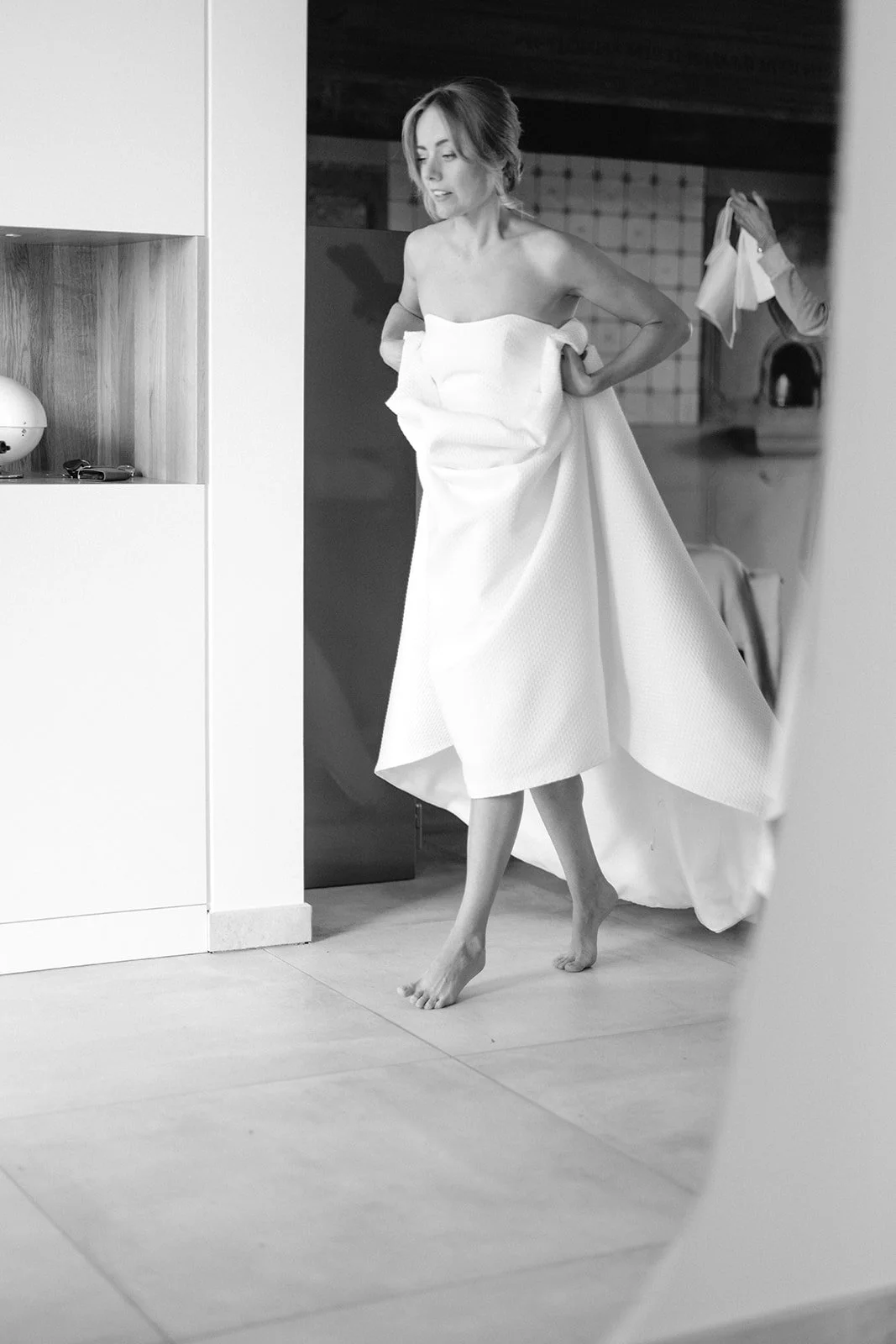 A woman in a wedding dress is putting on her gown, standing barefoot in a room with a mirror and kitchen background, seen through a reflection.