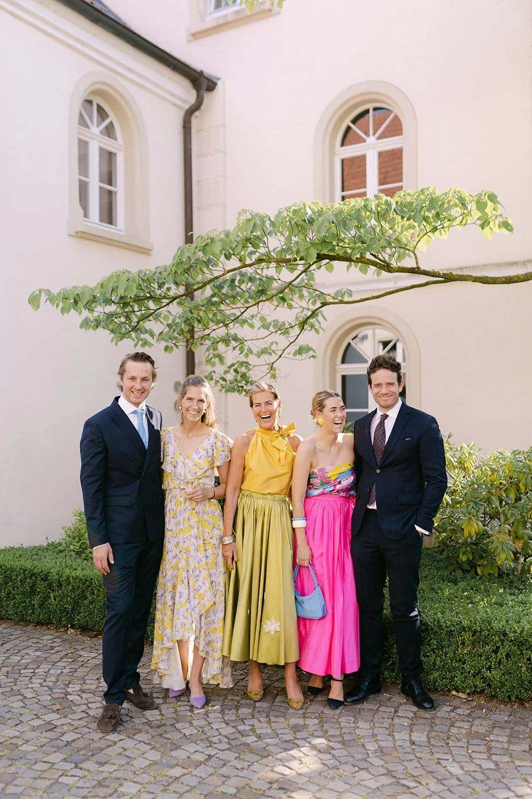 Group of five people standing outdoors on cobblestone path, smiling, with a cream-colored building and green foliage behind them. Four women and one man, dressed in colorful and formal attire.
