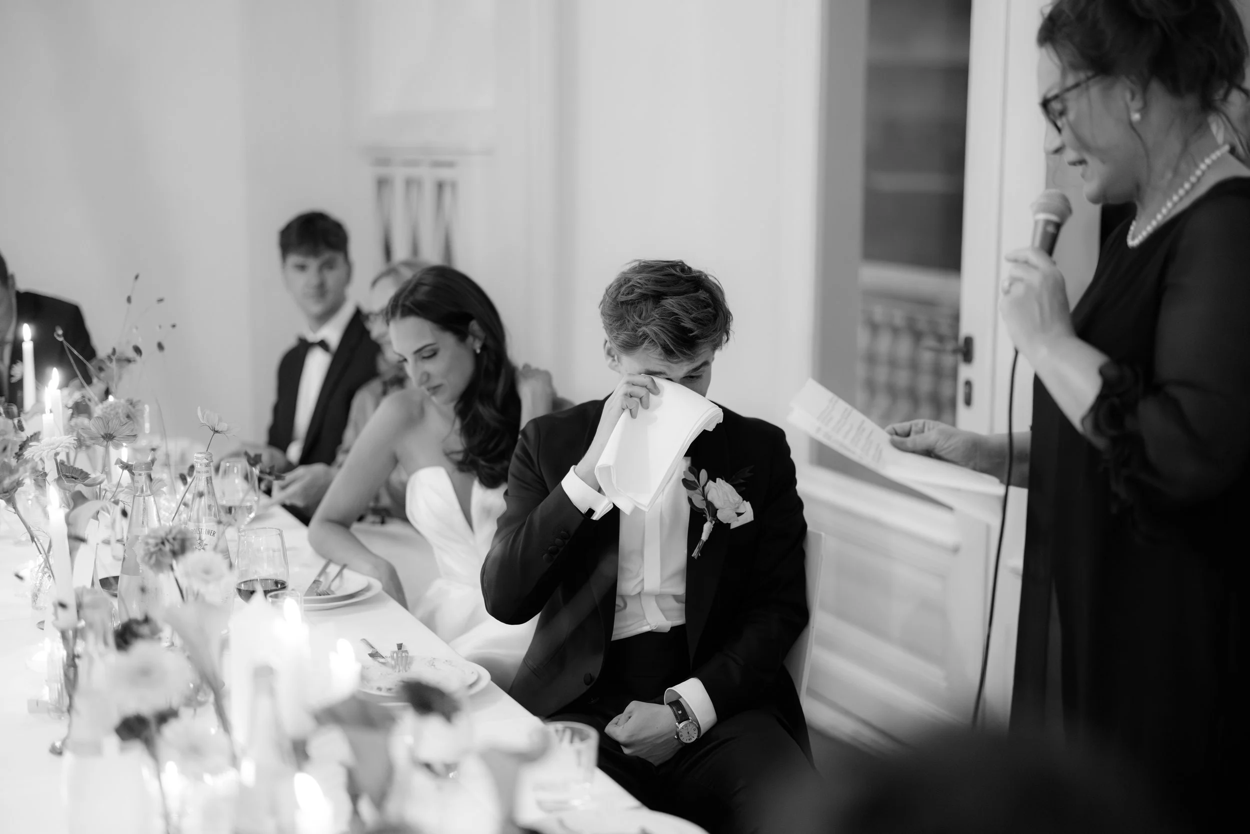 Black and white photo of a wedding reception with a bride, groom, and guests sitting at a decorated table. The groom appears emotional, wiping his eyes with a tissue while a woman speaker reads from a paper.